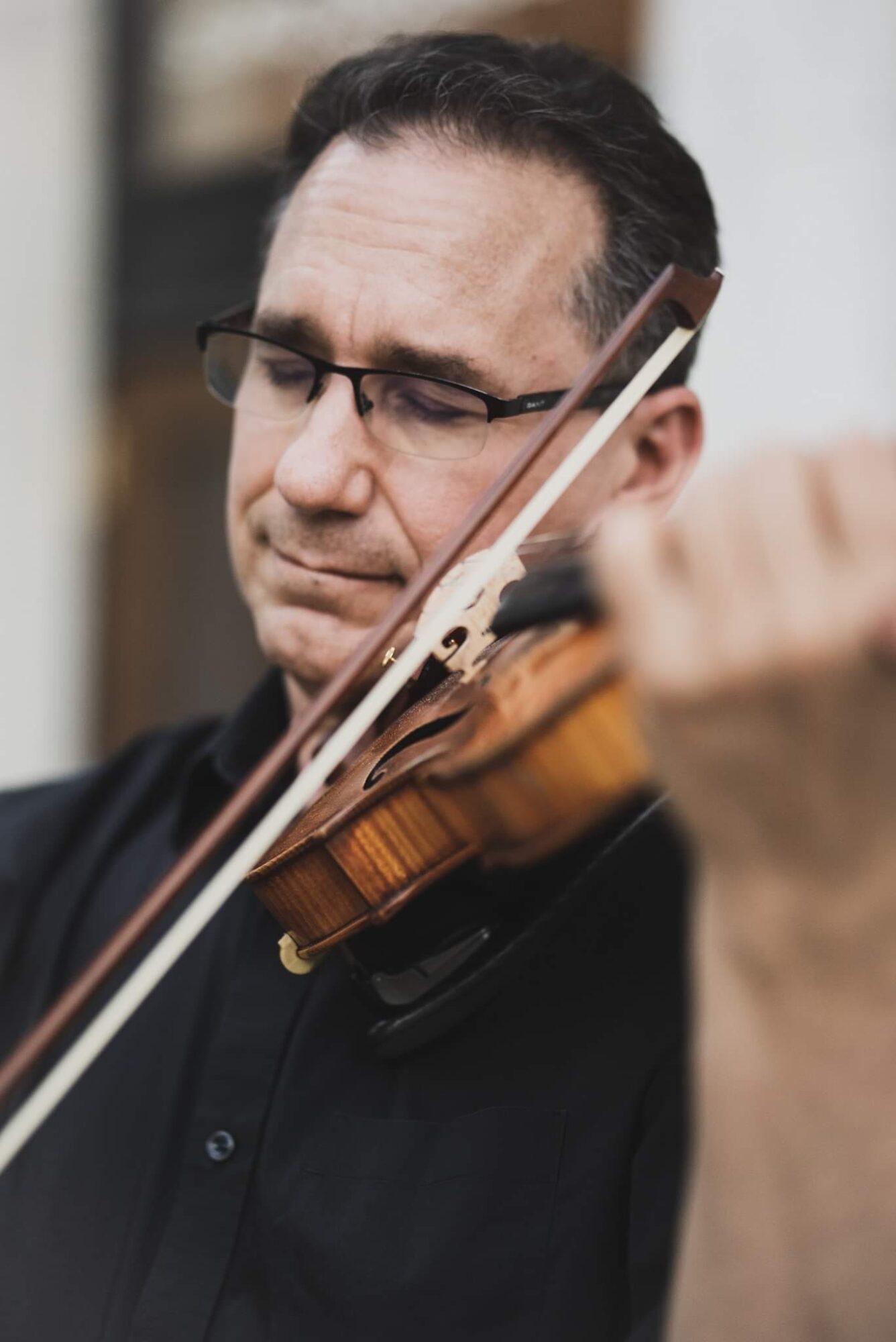 Man playing violin with glasses, focused expression, indoor setting, dark shirt, holding bow, close-up view.