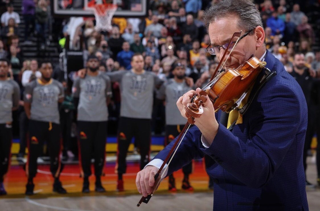 A man playing a violin in a sports arena with a basketball team in the background.