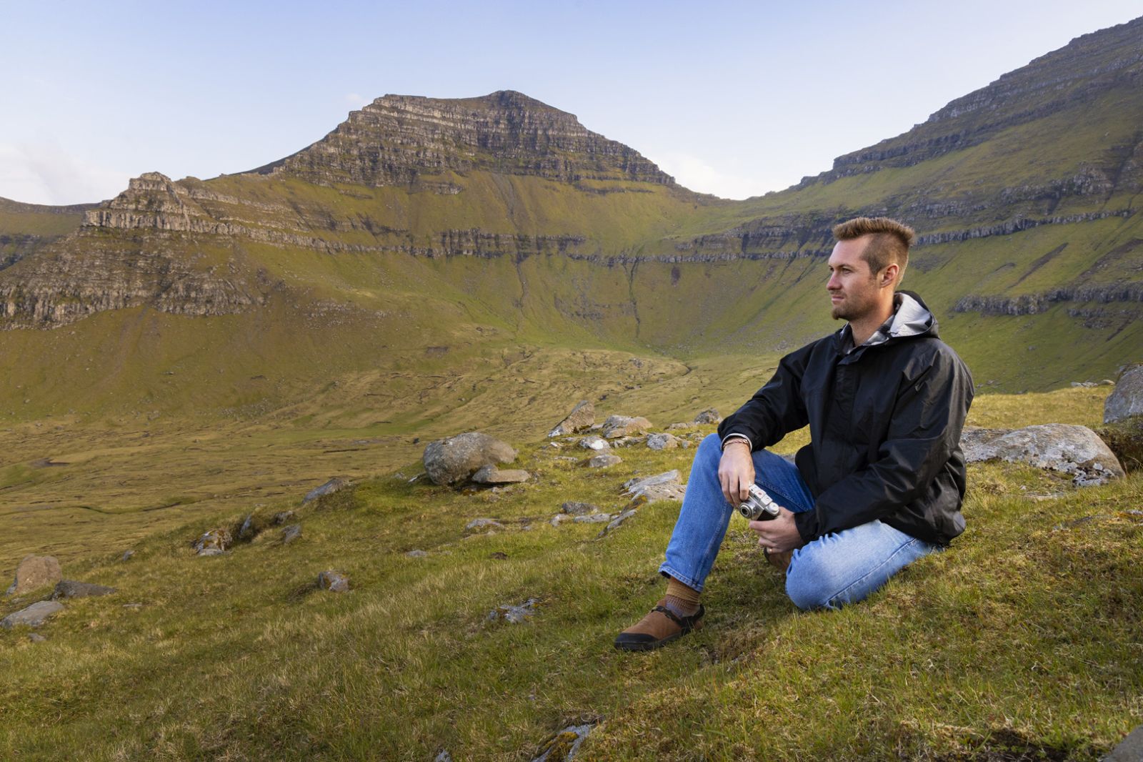 Man sitting on grass in a mountainous landscape with green hills and rocky formations, wearing a black jacket and blue jeans.
