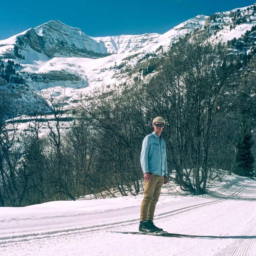 Person standing on snow with mountains and trees in background, clear sky, winter scene.