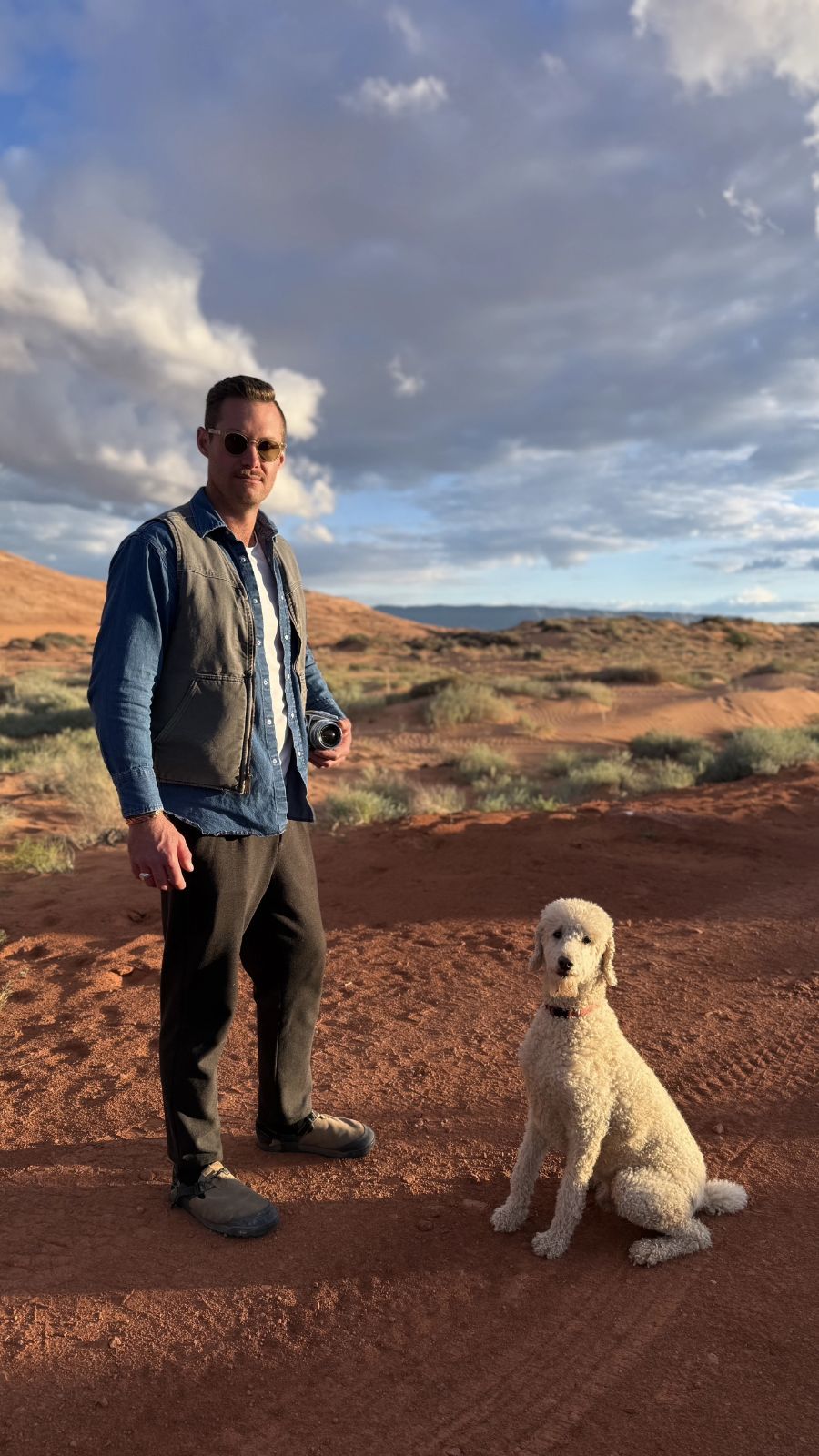 Man standing outdoors with a dog on a dirt path, cloudy sky above, landscape in the background.