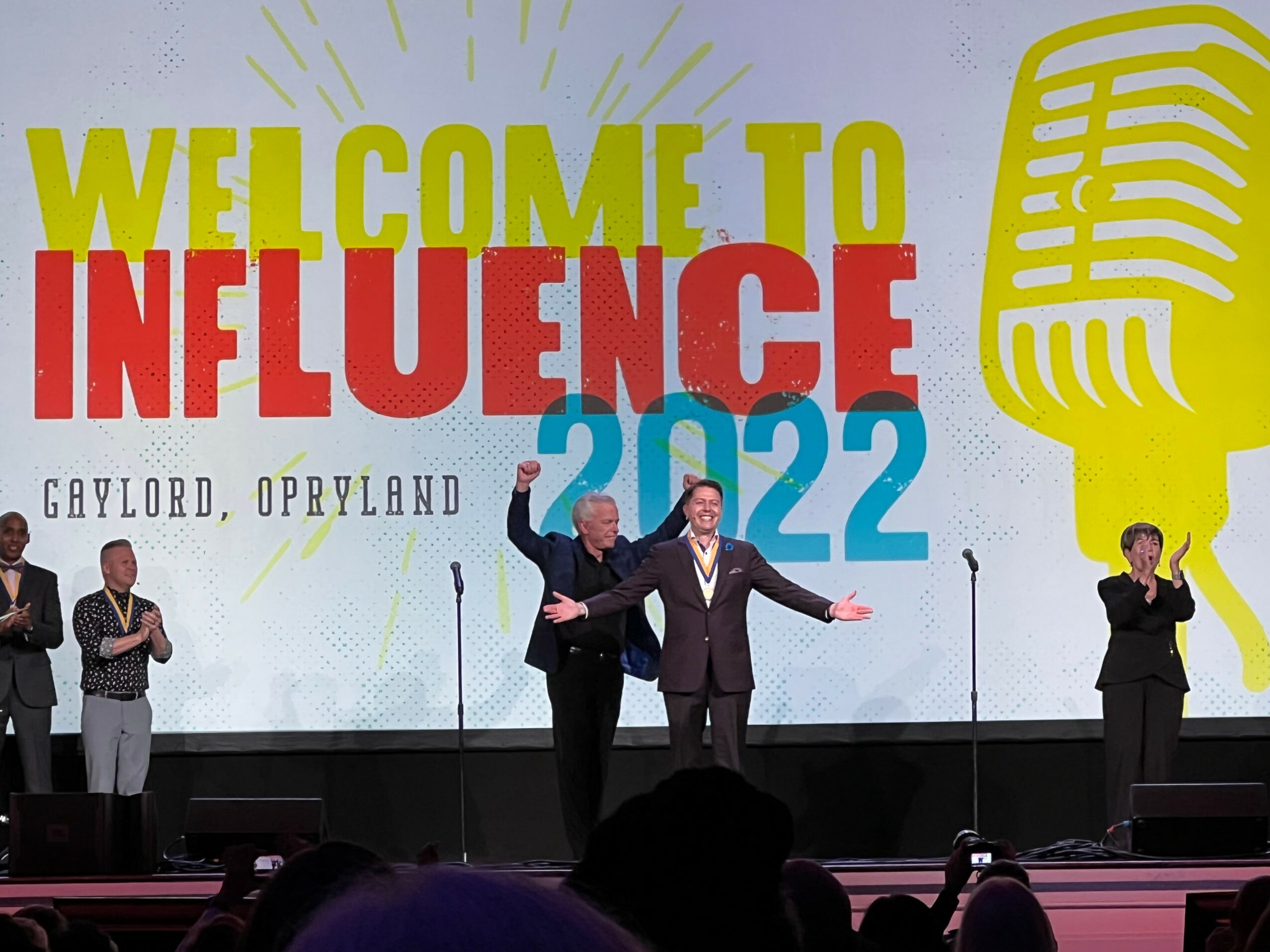 People on stage at a conference, with a large screen behind them displaying text and graphics, including a microphone icon.