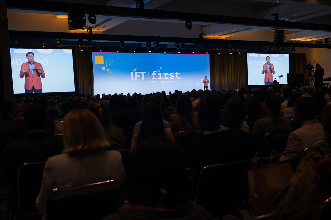 Large conference room with a stage, two screens showing a speaker, and an audience watching a presentation.