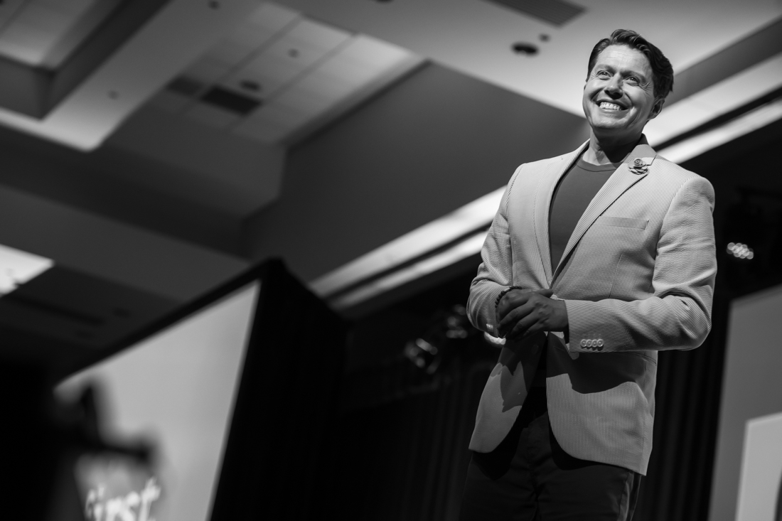 Smiling man in a suit standing on stage with a dark background, ceiling lights above, in black and white.
