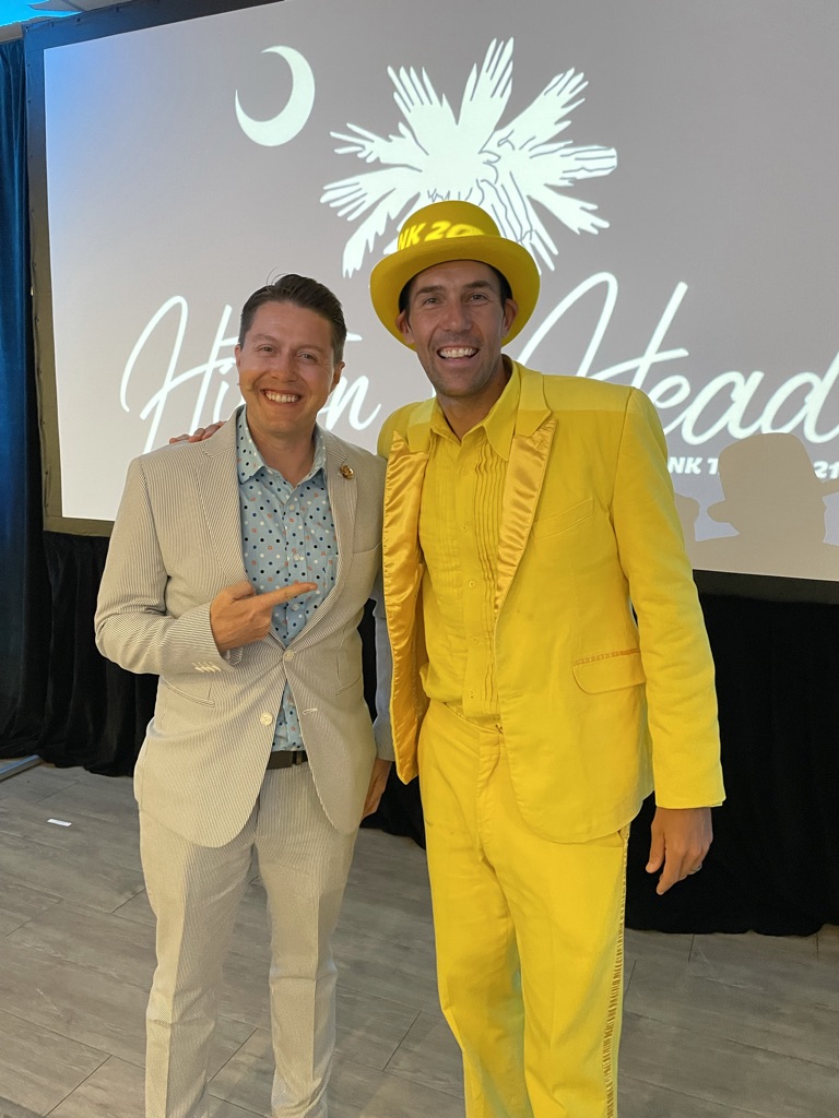 Two men in formal attire standing in front of a screen with a logo and text, one in a light suit, the other in a bright yellow suit and hat.
