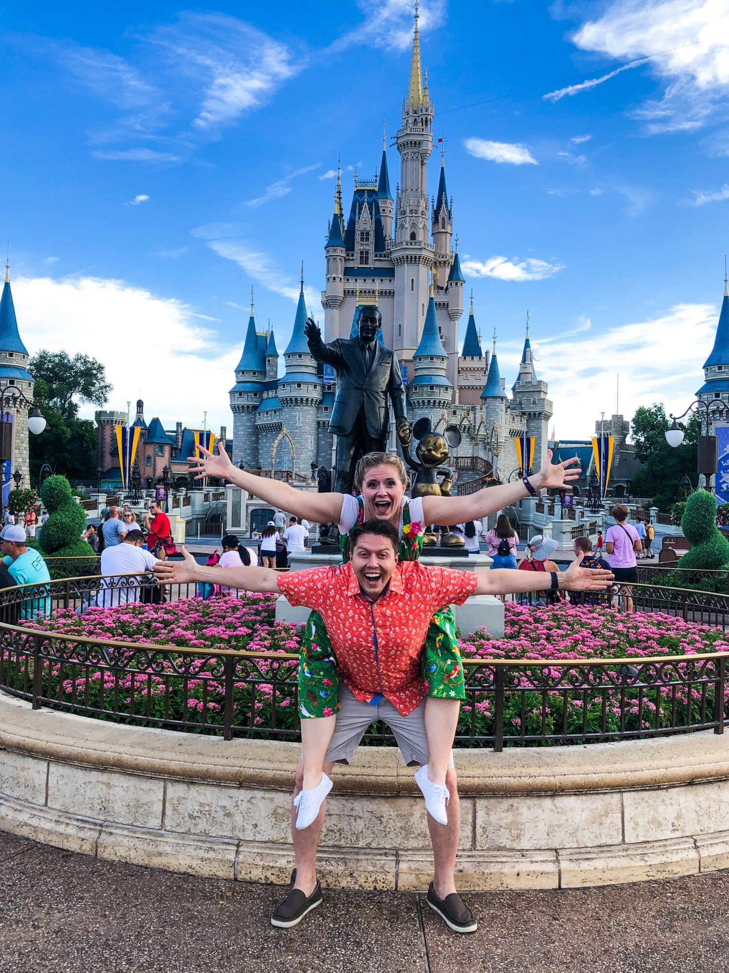 Two people pose in front of a castle at a theme park, with arms outstretched and smiling, surrounded by visitors and flowers.