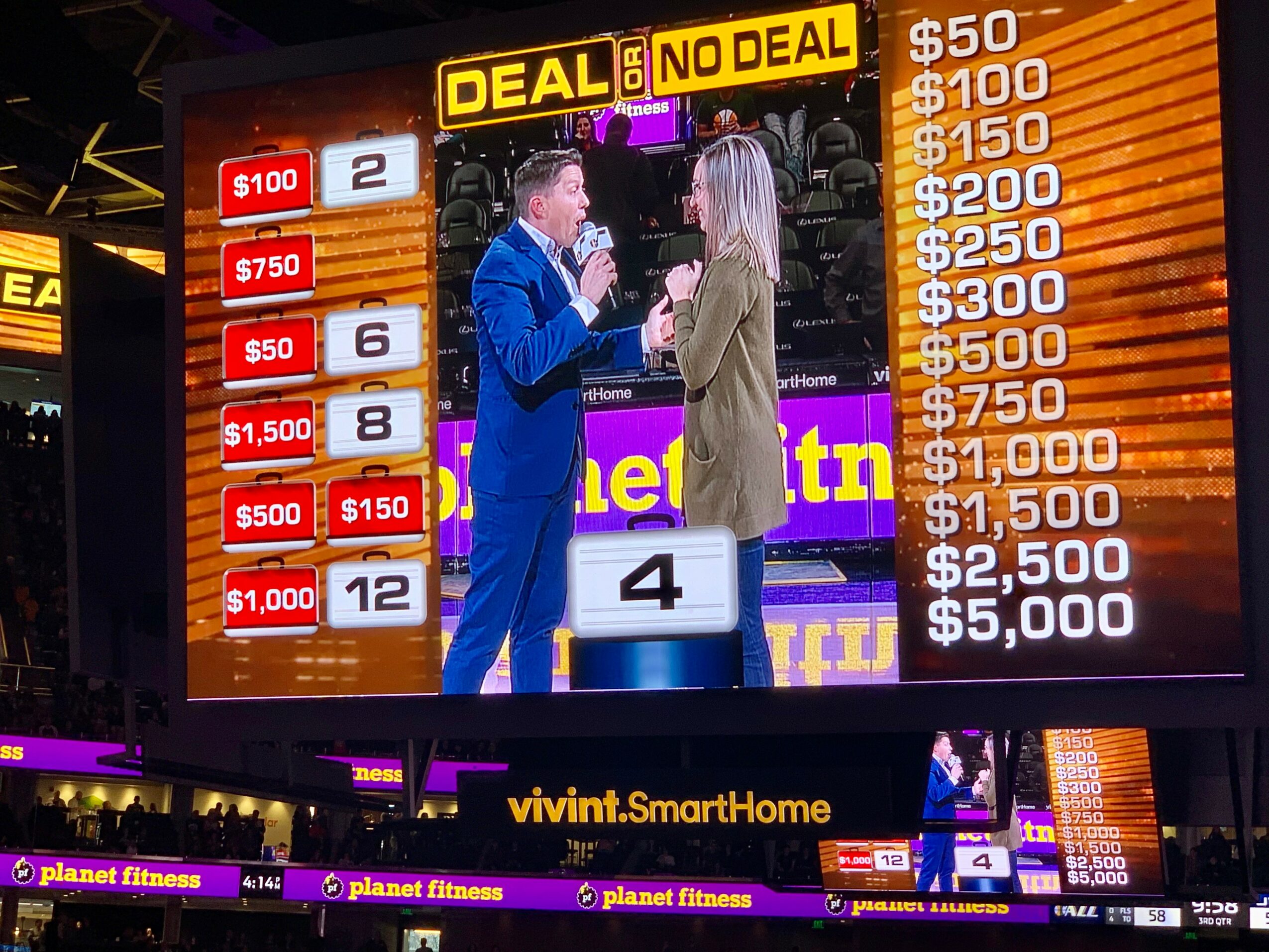 Two people stand on stage with a large screen behind them displaying numbers and text, including a dollar amount and a prize board.
