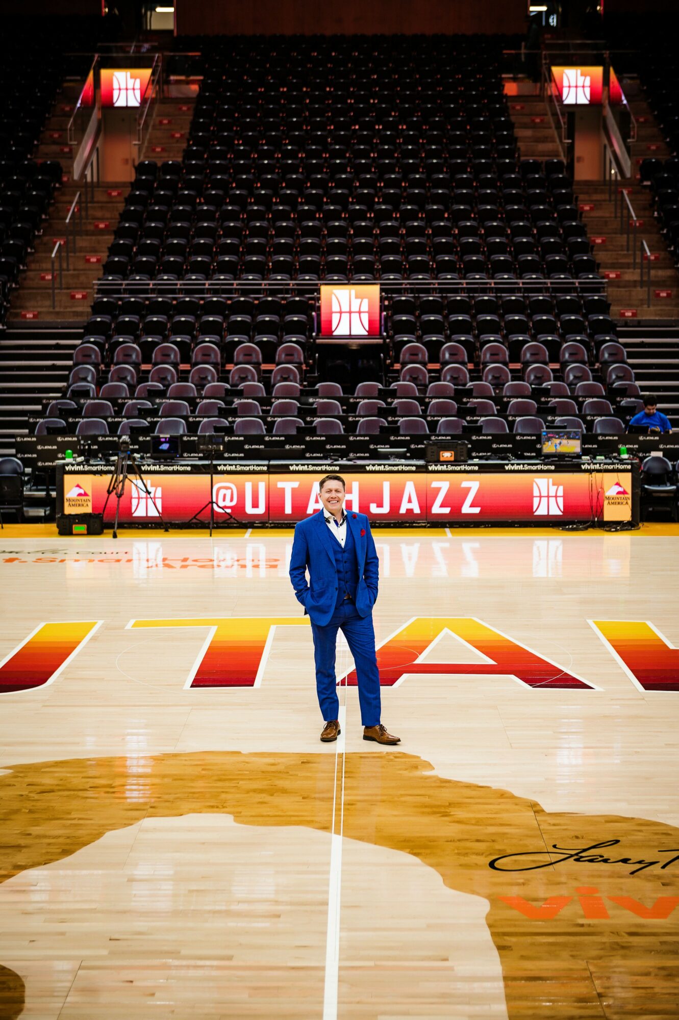 Man in blue suit standing on basketball court with empty seats in background, Utah Jazz signage visible.