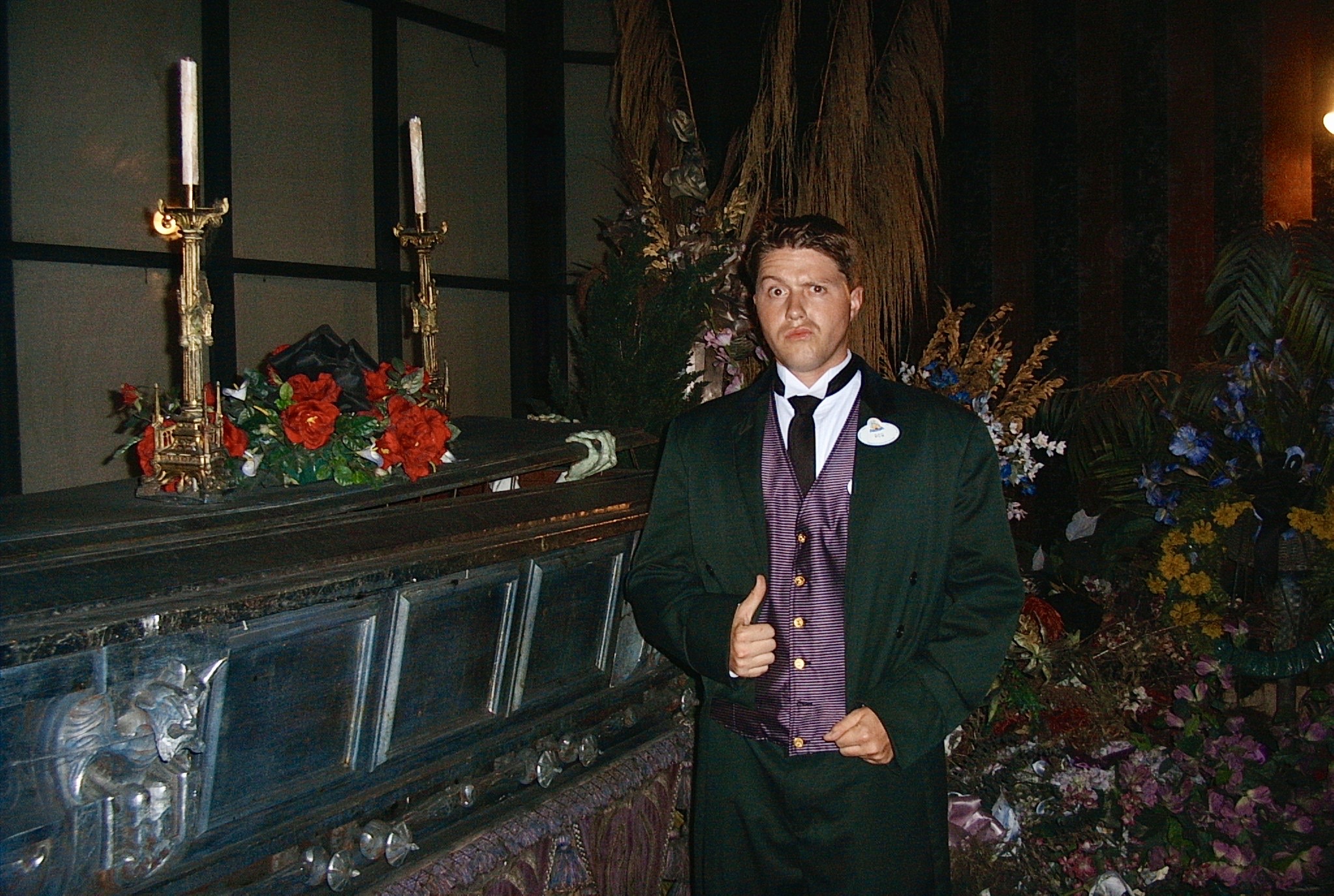 Young man in formal attire standing indoors with floral arrangements and candles in the background.