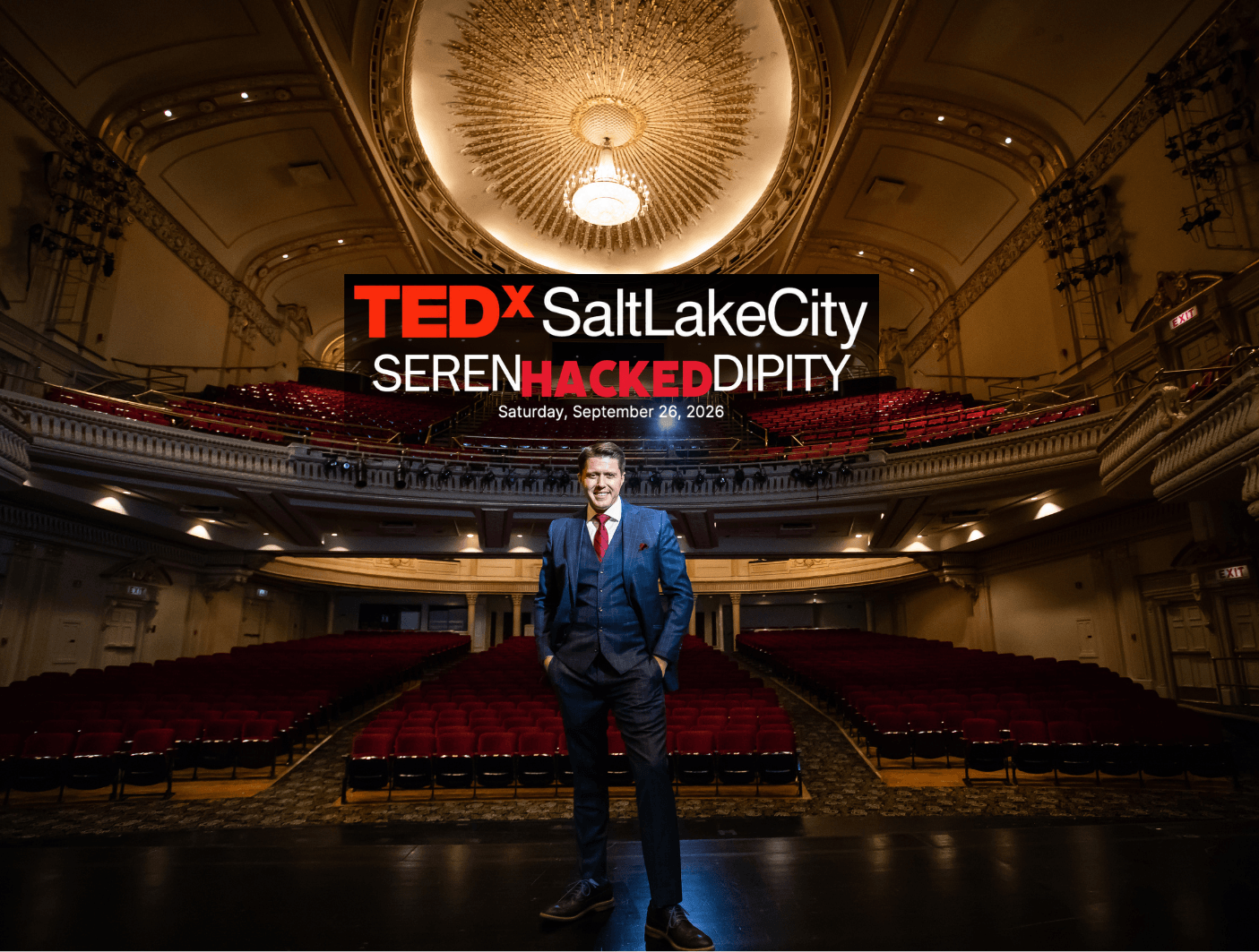 Man in suit standing on stage in an ornate theater with a large chandelier, TEDxSaltLakeCity sign above him, empty seats around.