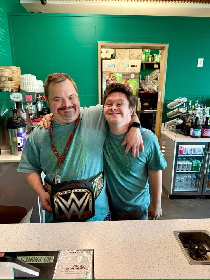Two smiling men with arms around each other in a store, one wearing a WWE championship belt, green wall background.