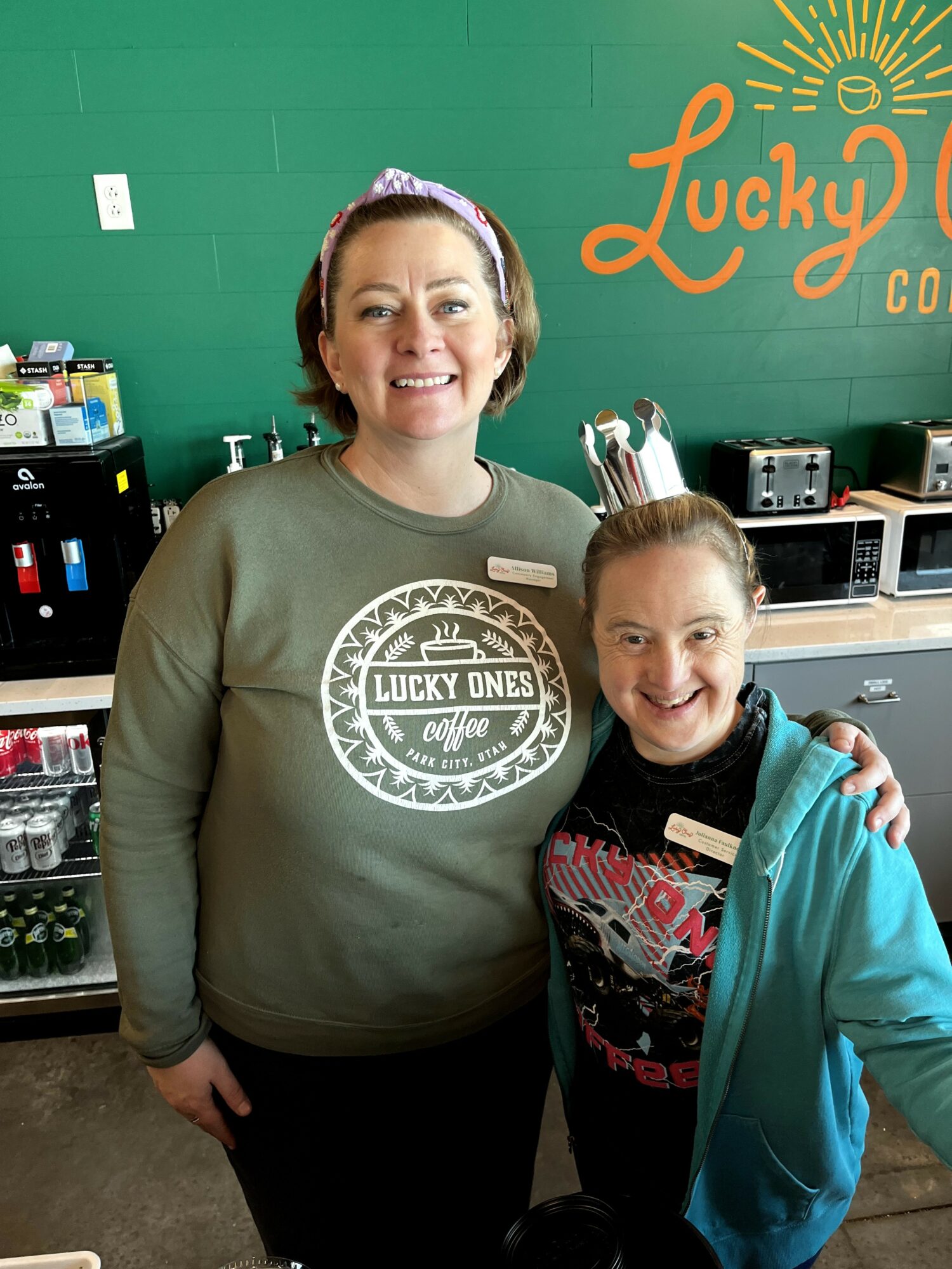 Two women smiling in a store, one wearing a green shirt with a logo, the other in a blue jacket, with a green wall and store items behind them.