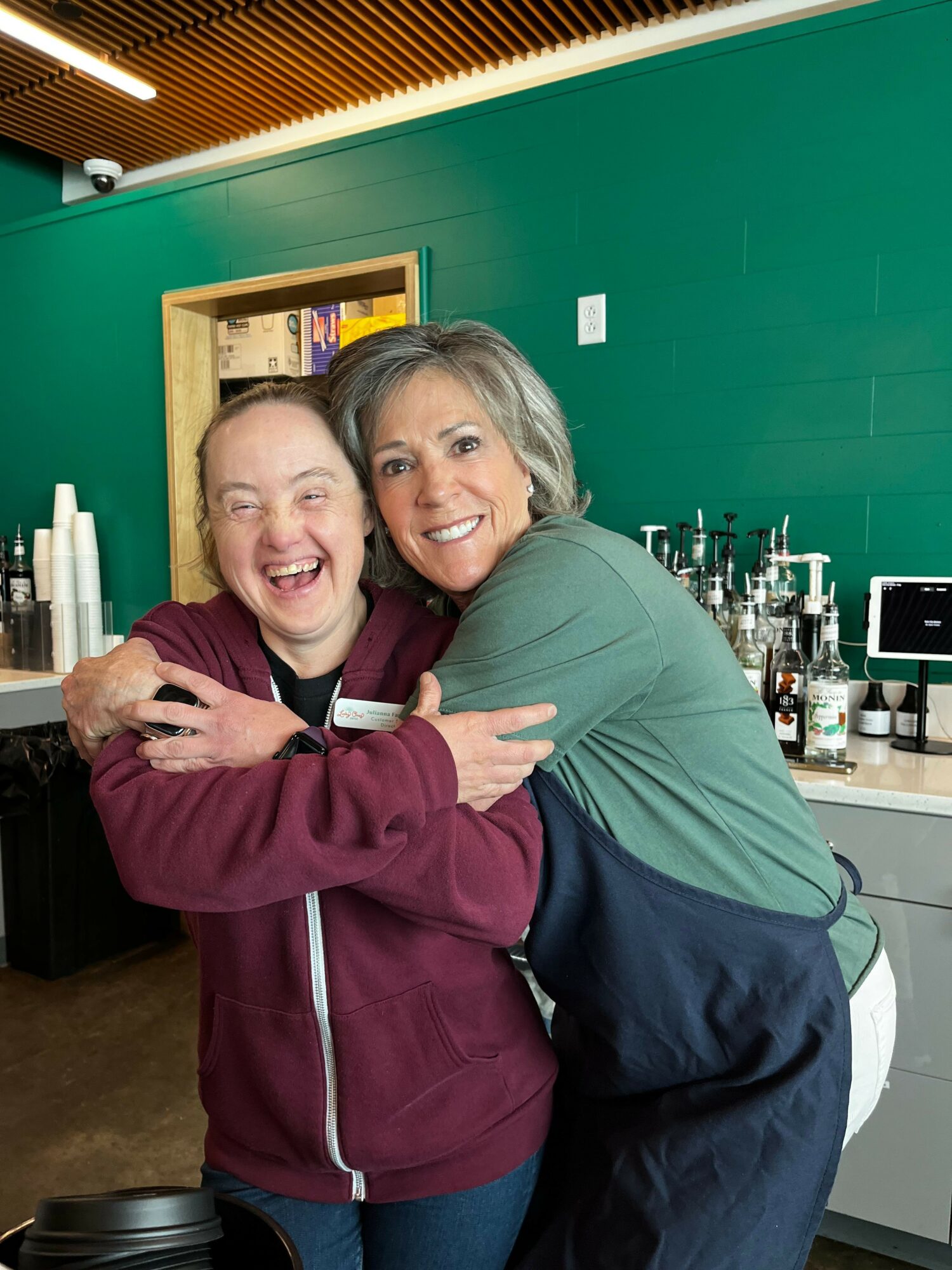 Two women hugging and smiling in a room with green walls and equipment in the background.