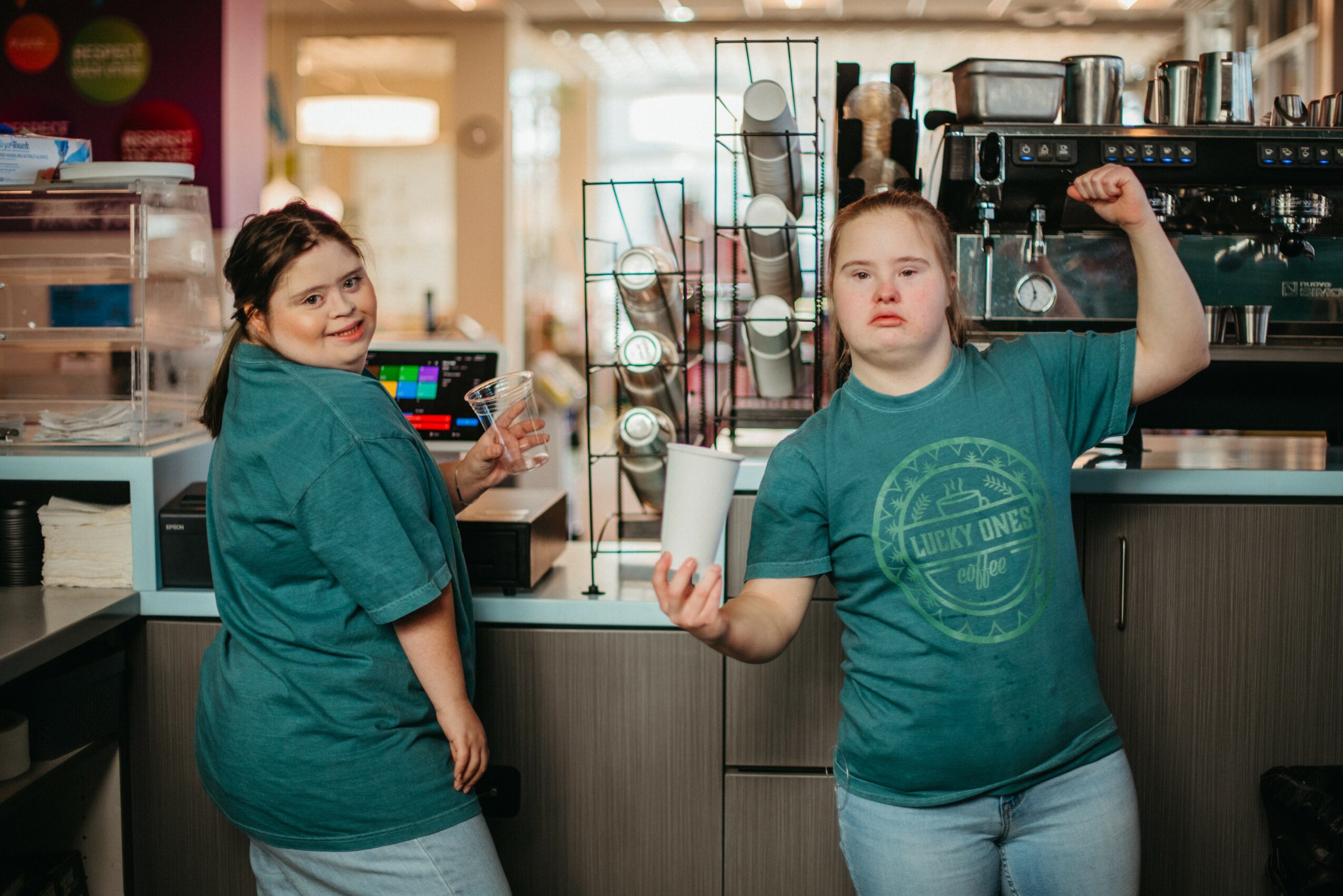 Two girls in a cafe, one smiling and the other flexing her arm, standing behind a counter with coffee equipment.