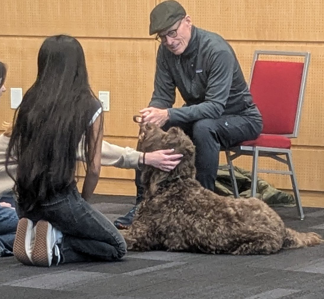 Person with long hair and a person with a cap pet a large, curly-haired dog on the floor in a room.