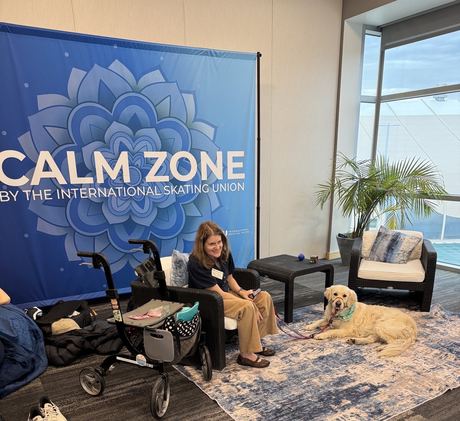 Woman sitting in a chair with a service dog lying on the floor nearby, in a room with a large window and a blue banner that reads 'Calm Zone'.