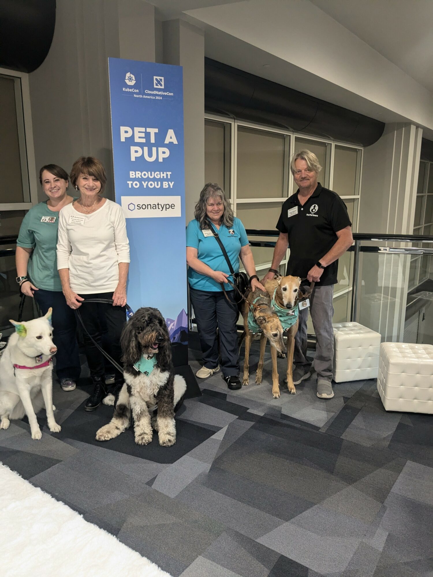 Four people with three dogs standing next to a blue sign that reads 'PET A PUP' at an indoor event.