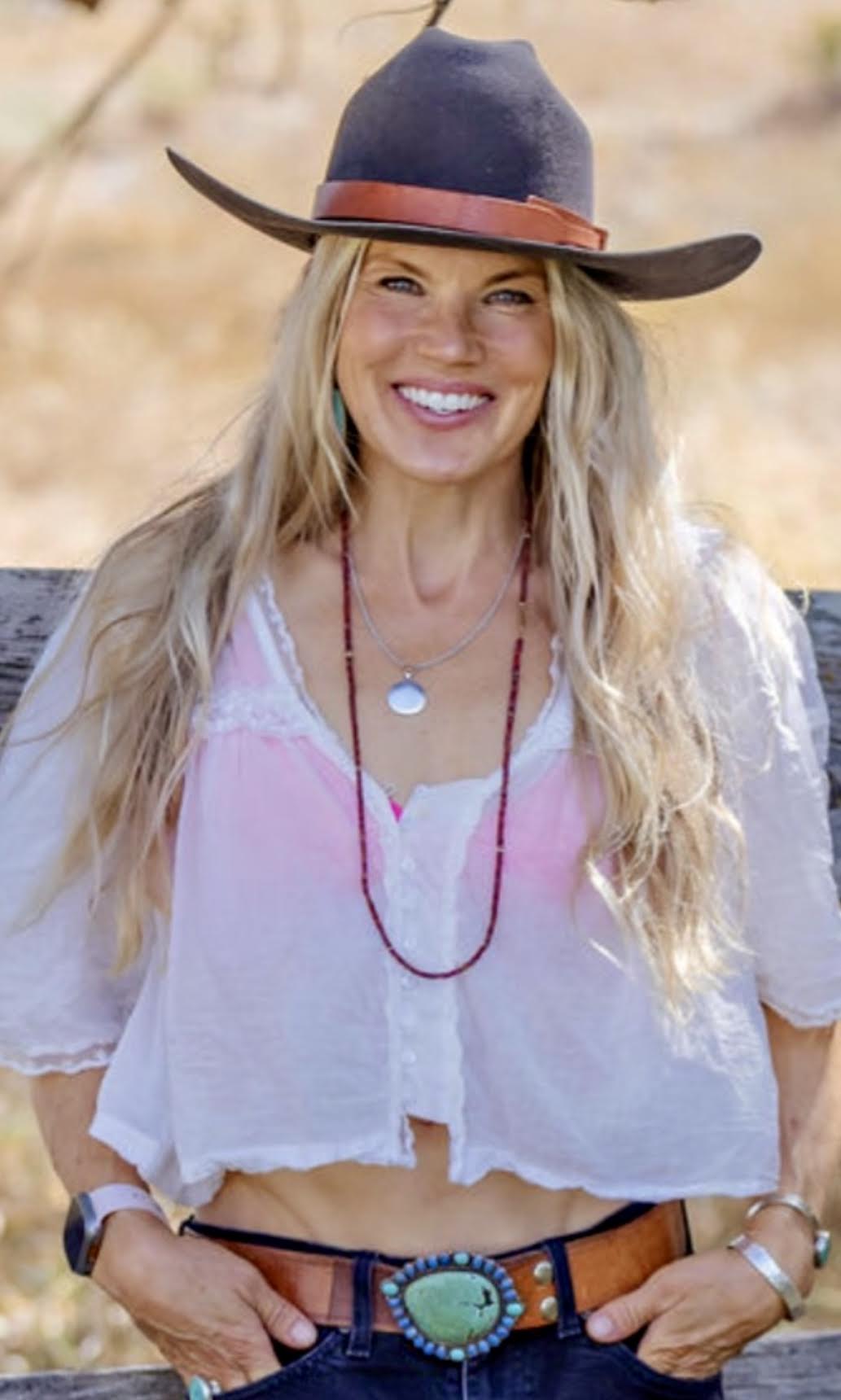 Woman smiling outdoors wearing a wide-brimmed hat, white blouse, and jewelry.