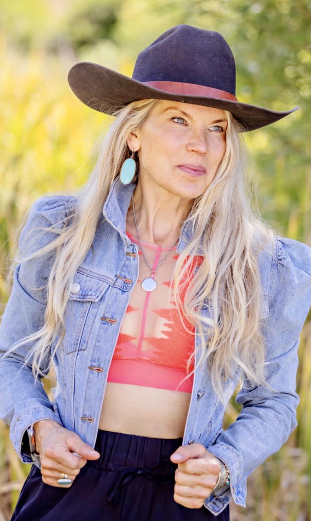 Woman with long blonde hair wearing a wide-brimmed hat, denim jacket, and colorful top outdoors.