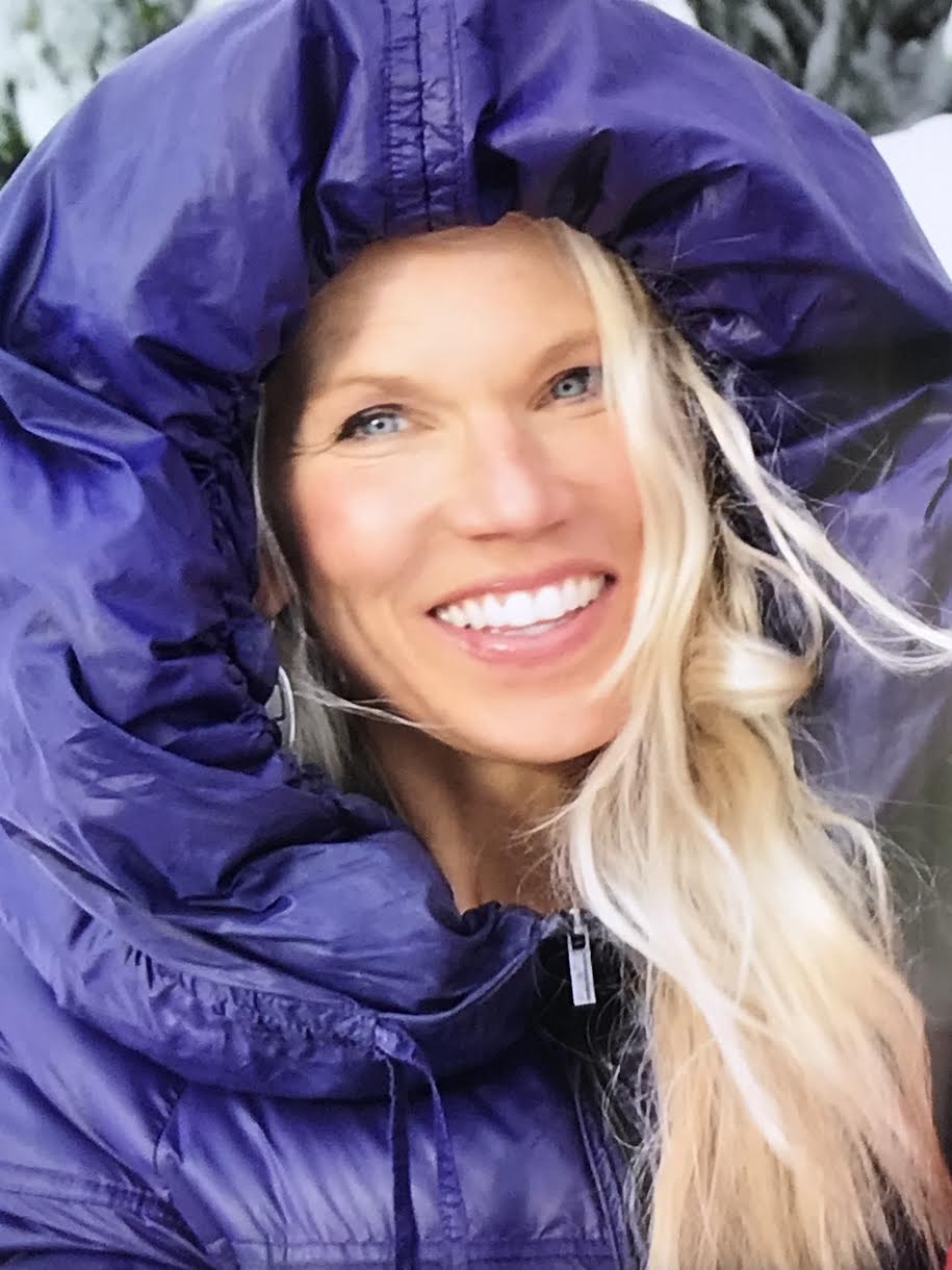 Woman smiling with blonde hair, wearing a purple jacket with hood over her head, outdoors.