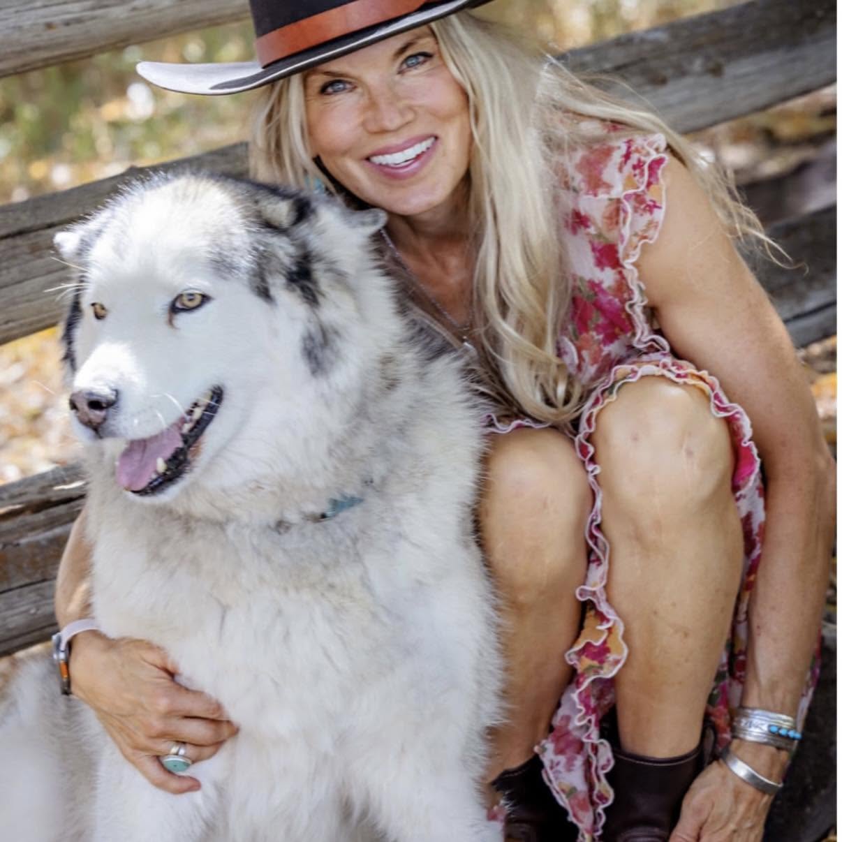 Woman with long blonde hair wearing a hat and floral dress, smiling with a large Siberian Husky outdoors.