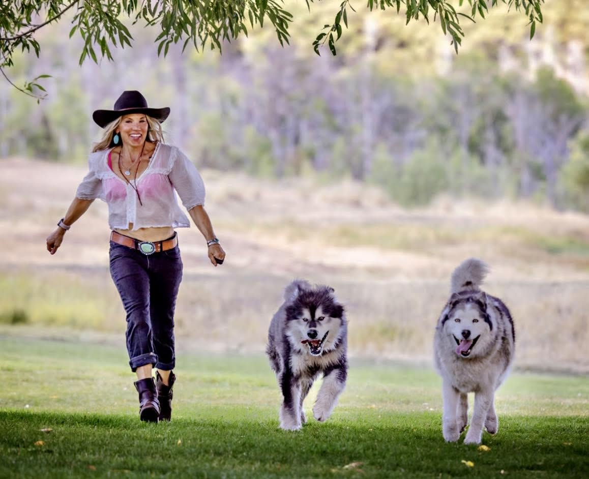 Woman walking with two dogs in a grassy park, trees in background, sunny day.