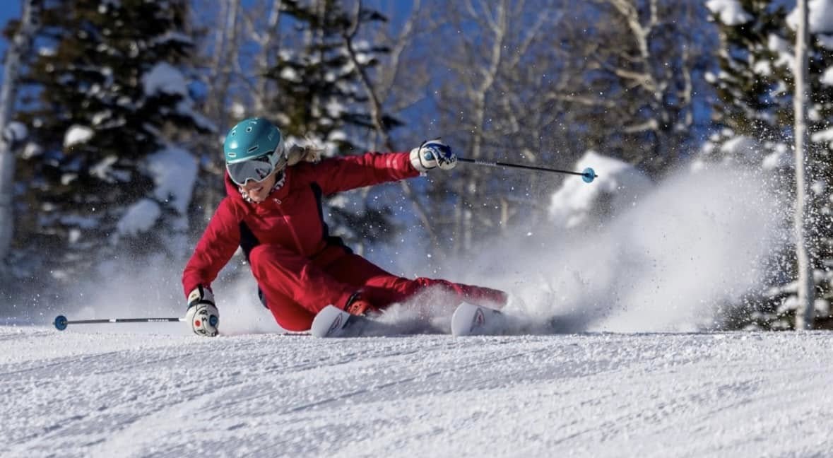 Person skiing downhill in a snowy landscape with trees, wearing a red outfit and teal helmet, creating snow spray.