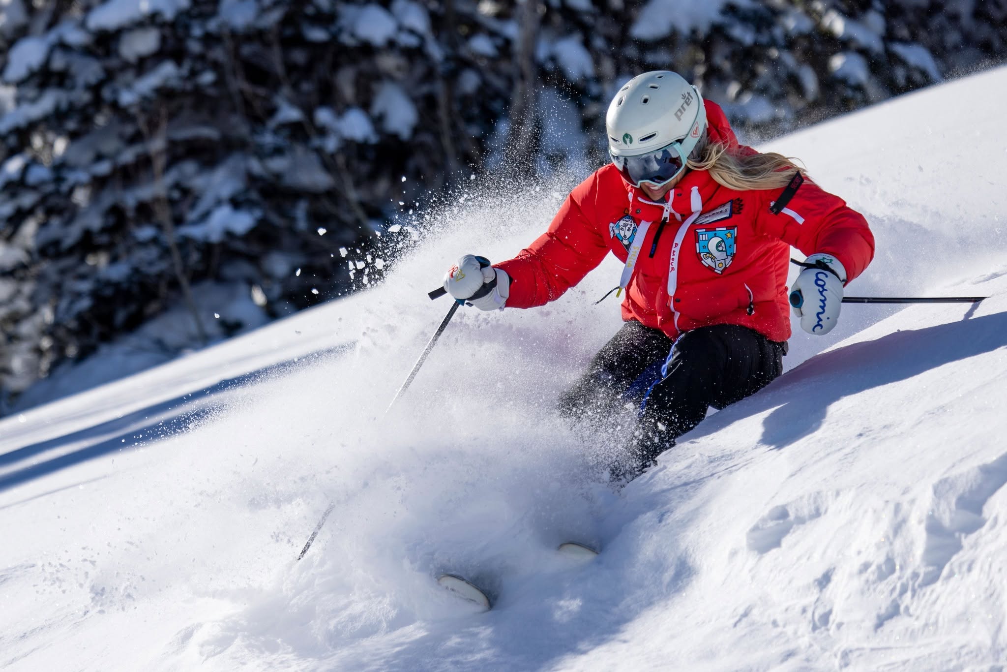 Person skiing downhill in snow, wearing a red jacket, white helmet, and goggles, surrounded by snow-covered trees.