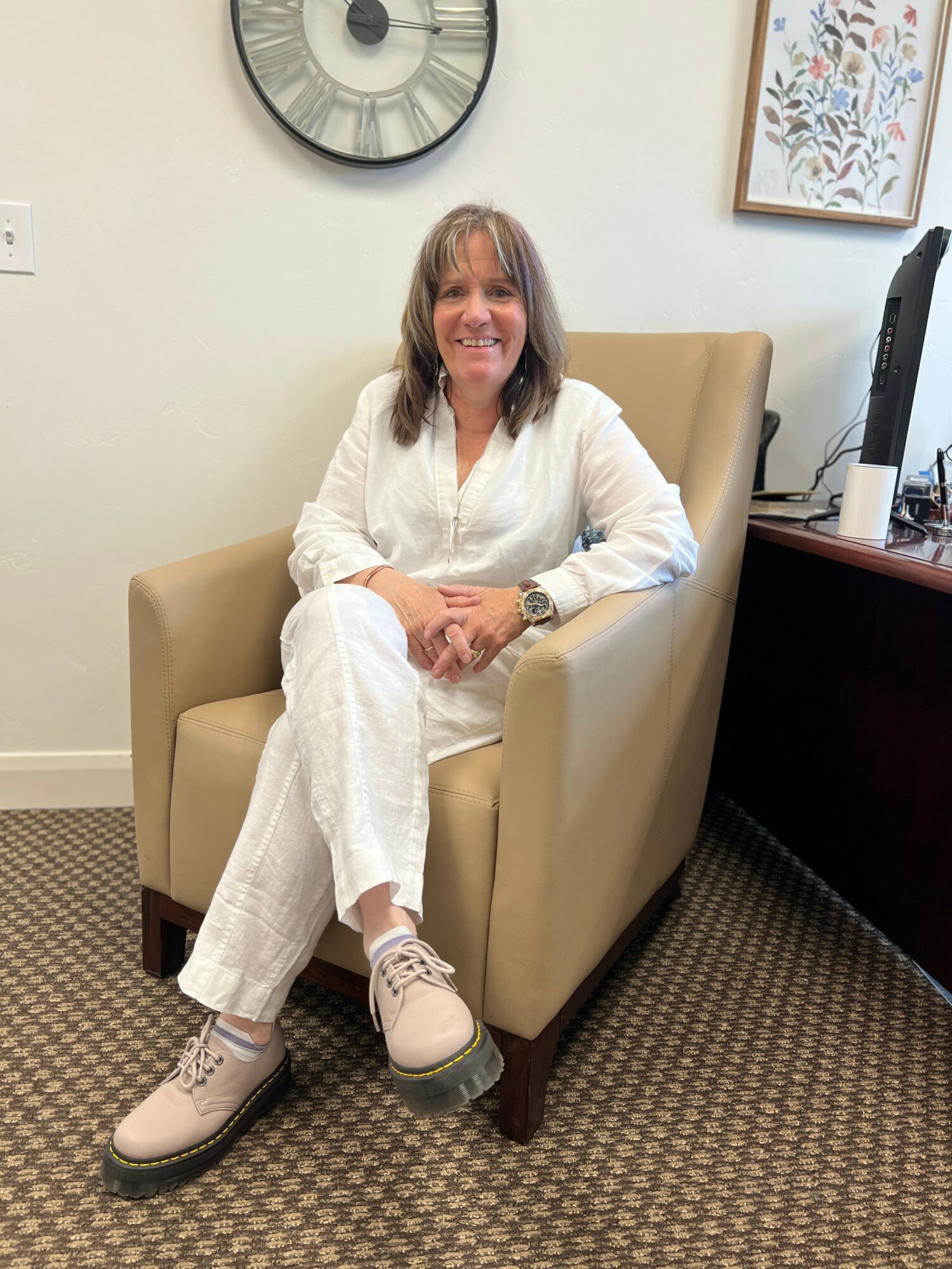 Woman sitting in a beige armchair, smiling, in an office with wall clock and framed artwork, wearing white outfit and beige shoes.