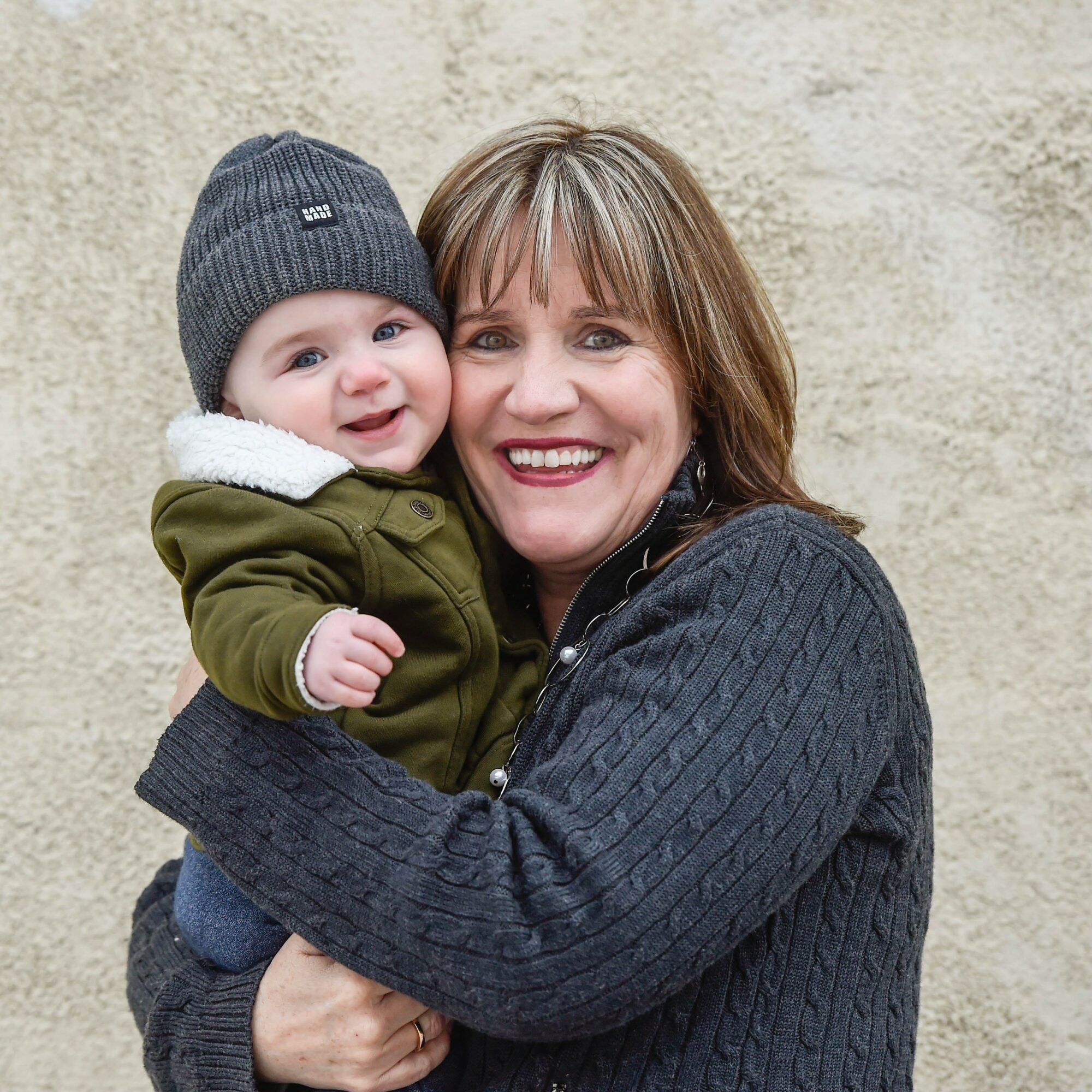 Woman holding smiling baby outdoors against a beige wall, both dressed warmly, woman with short brown hair, baby in a beanie and jacket.