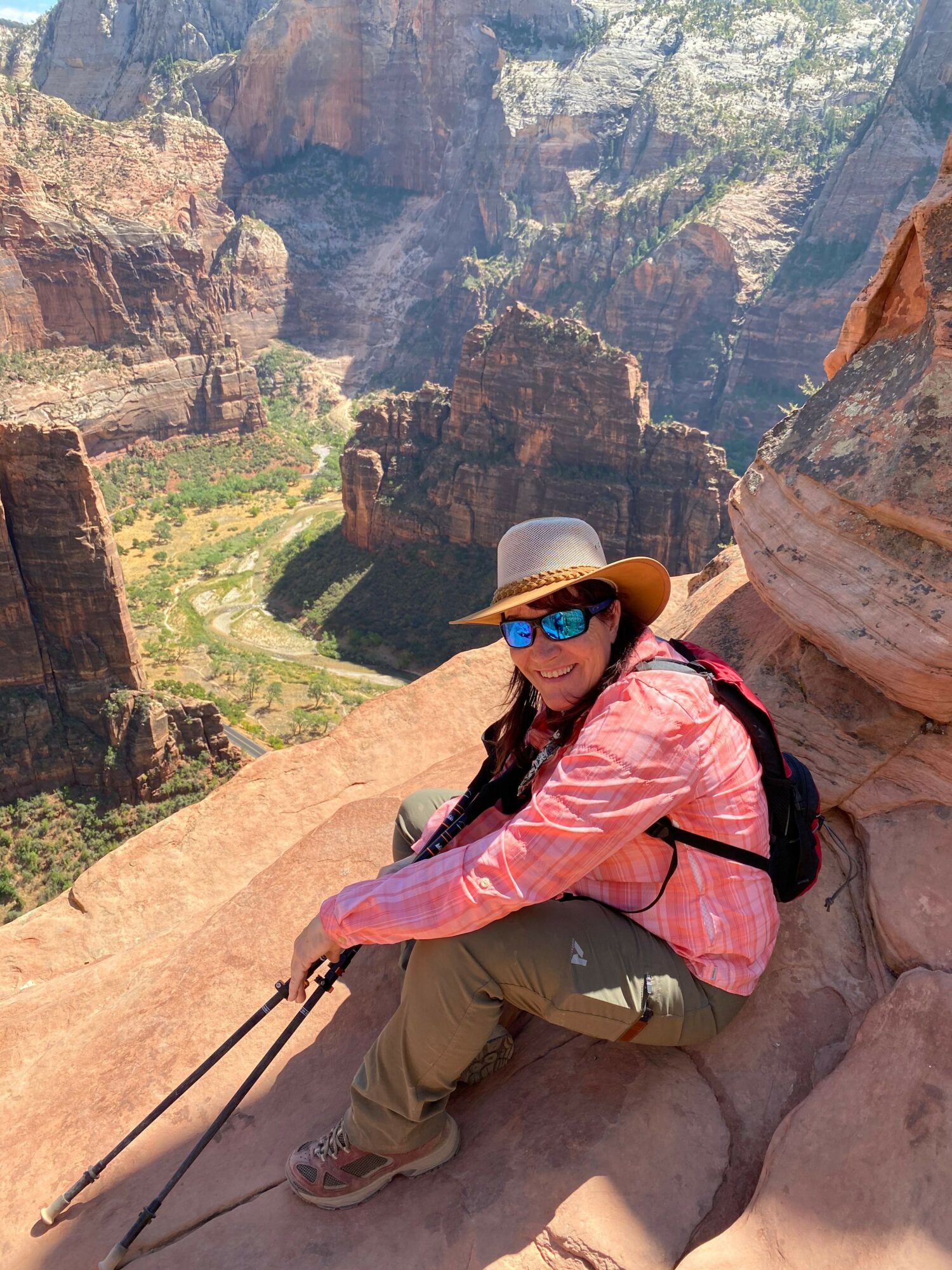 Person wearing a hat, sunglasses, pink jacket, and khaki pants, sitting on a rock ledge with a canyon view.