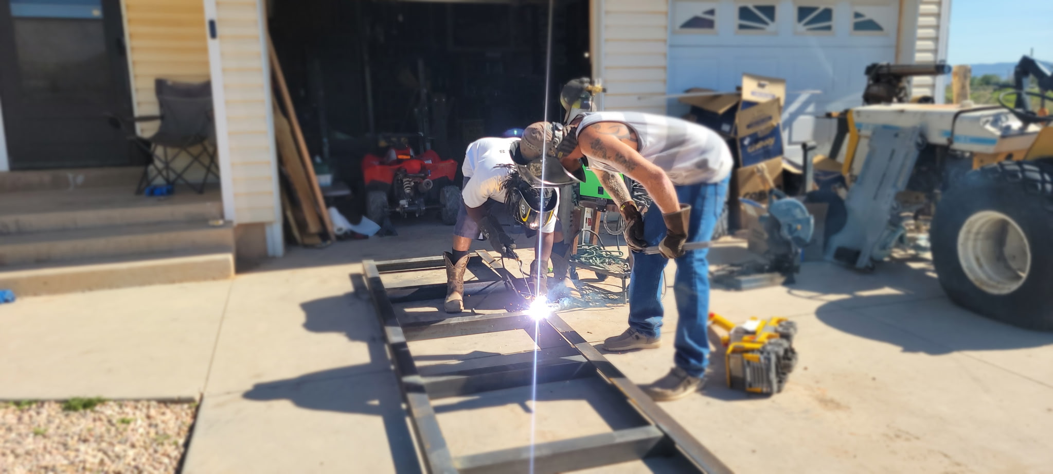 Two people welding metal frame outdoors, sparks flying, with tools and equipment nearby, in a sunny setting.
