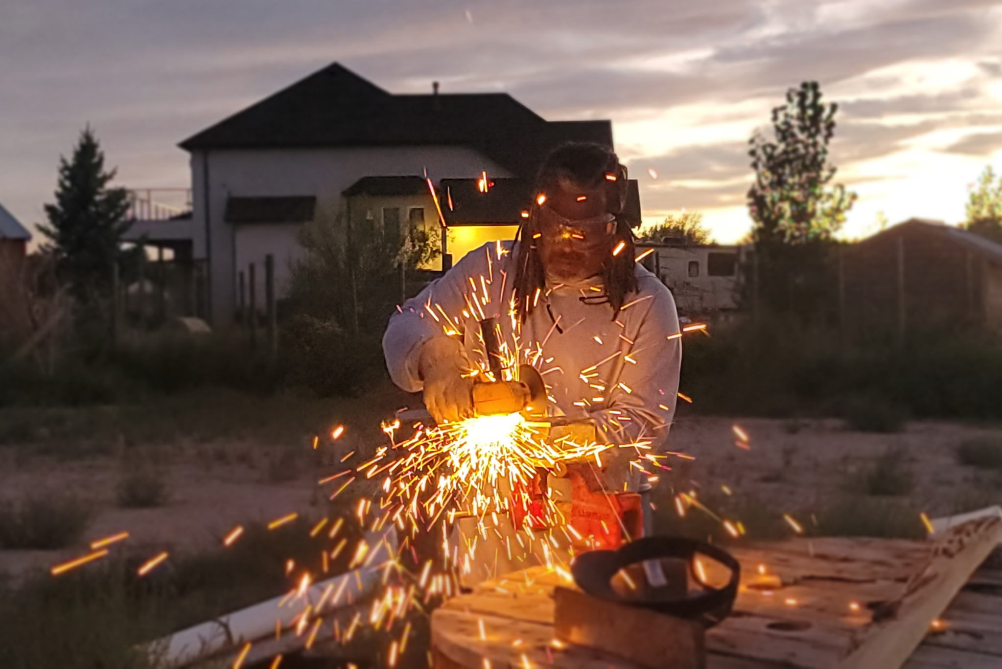Person welding outdoors with sparks flying, house and trees in background at sunset, wearing protective gear.