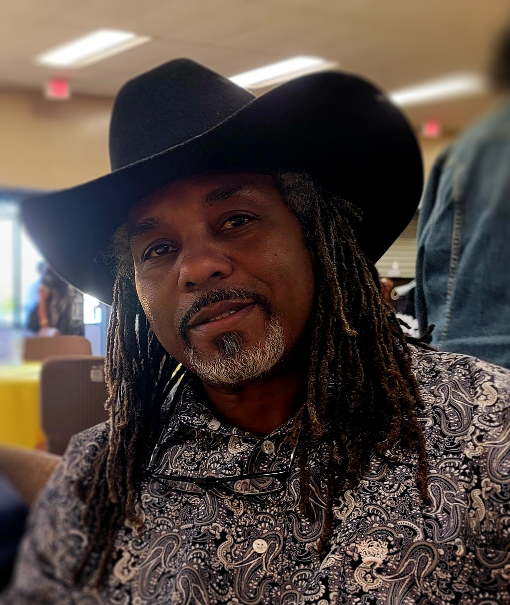 Man with long dreadlocks wearing a large black hat and patterned shirt, indoors with blurred background.
