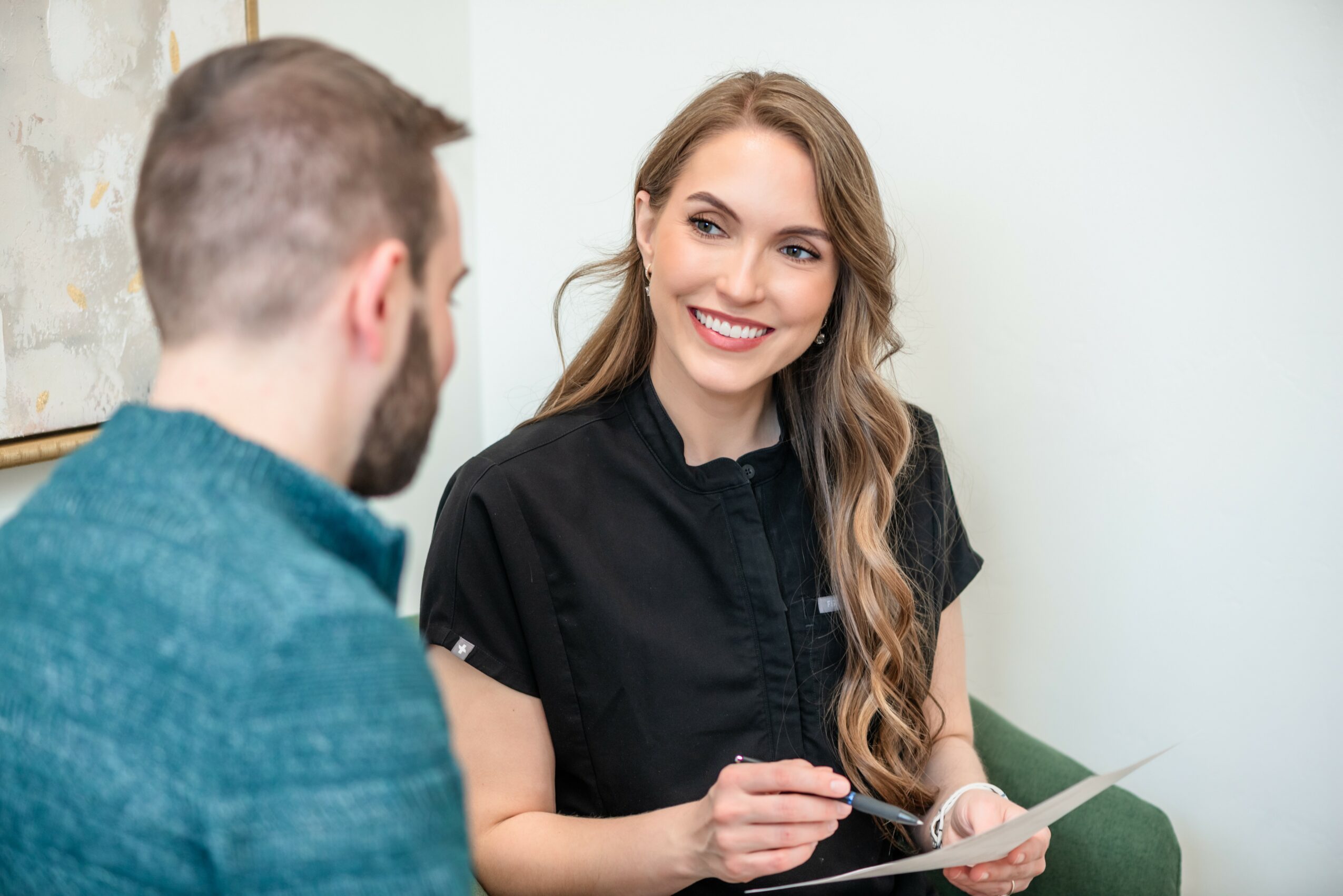 A woman with long wavy hair smiling and holding a pen, talking to a man with short hair and a beard, in a bright room.