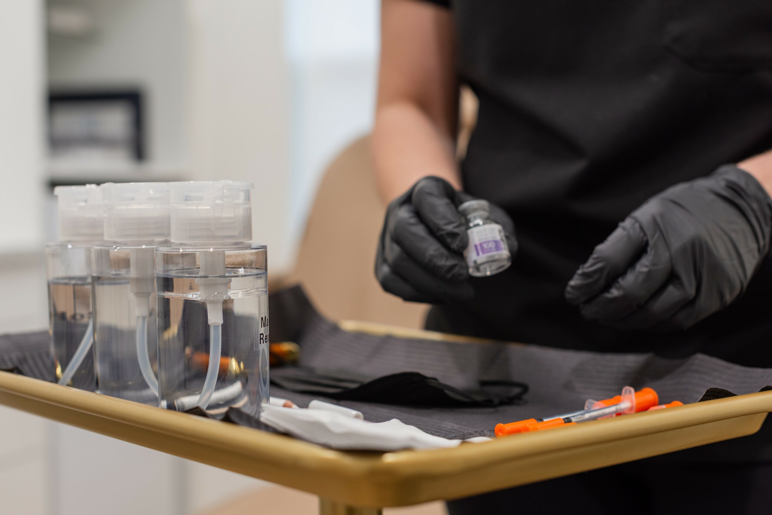 Person wearing black gloves holding a small vial, with laboratory equipment on a table, background blurred.