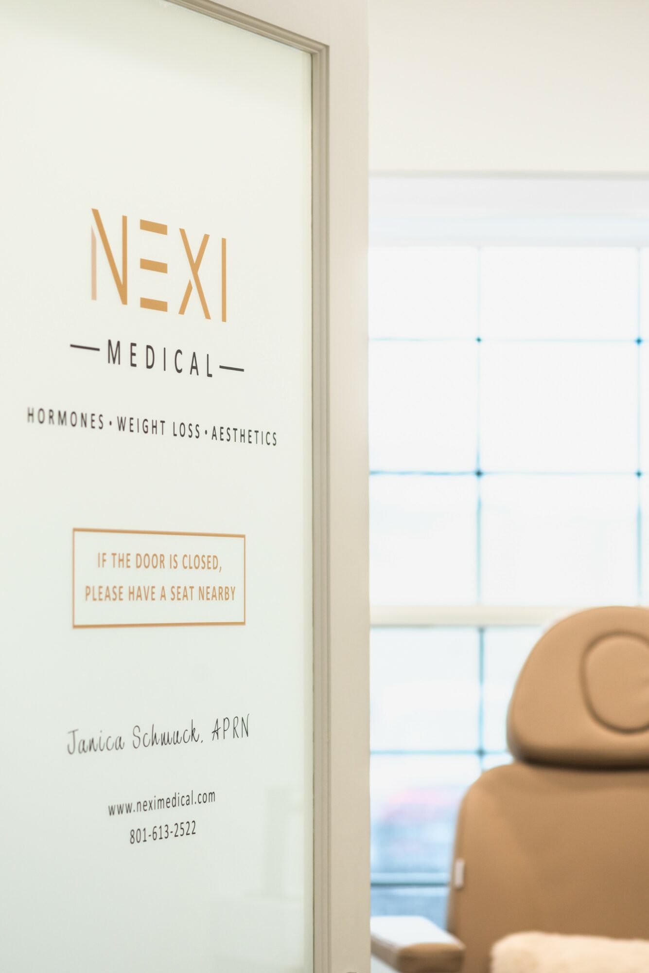 Medical clinic door with text, and a beige chair in a bright room with large windows.