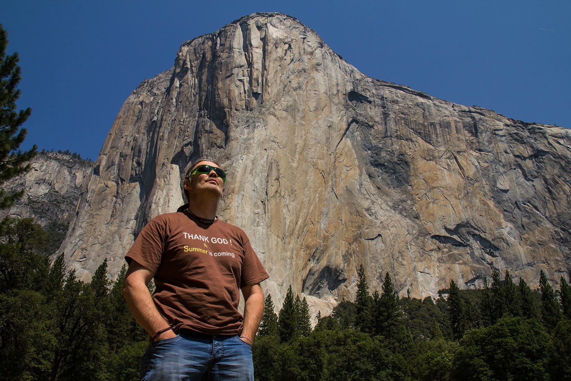 Person standing outdoors with a large mountain in the background, wearing sunglasses and a brown T-shirt, trees at the base.