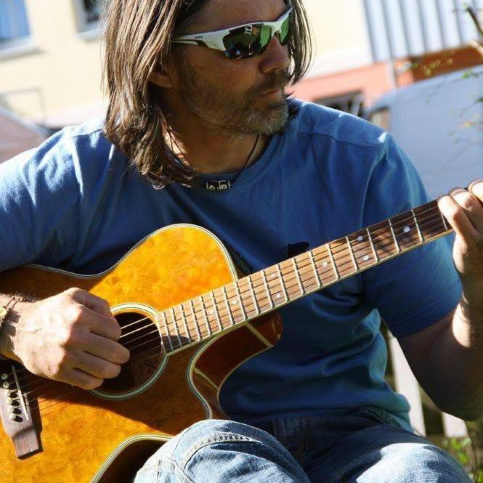 Man with long hair, sunglasses, blue shirt, playing an acoustic guitar outdoors, sunlight, buildings in background.