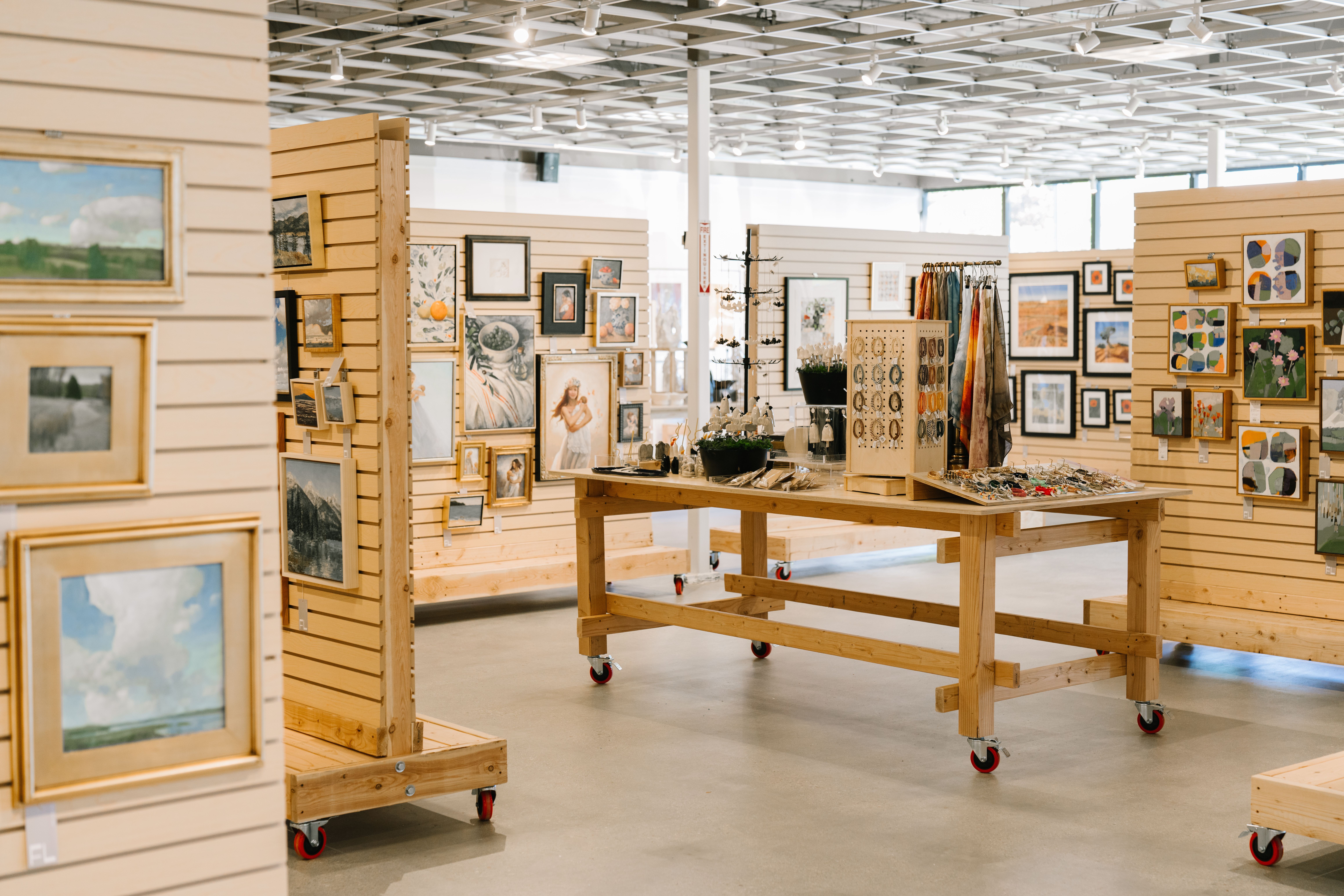 Art gallery with framed paintings and artwork displayed on wooden partitions and walls, with a central table and bright lighting.