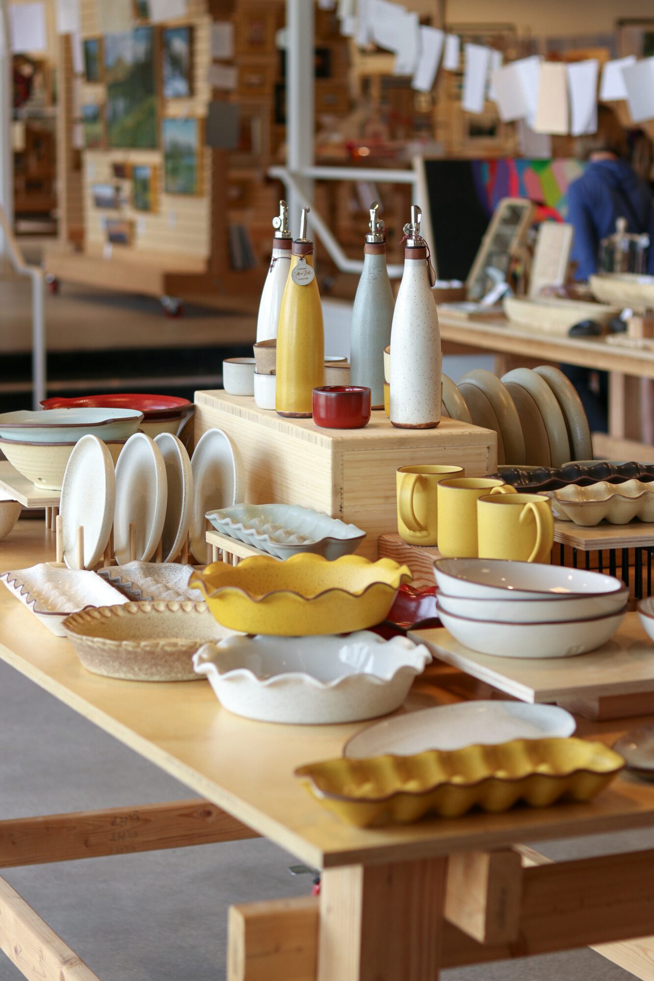 Display of various ceramic dishes, bowls, and mugs on a wooden table at a market stall, with bottles and a person in background.