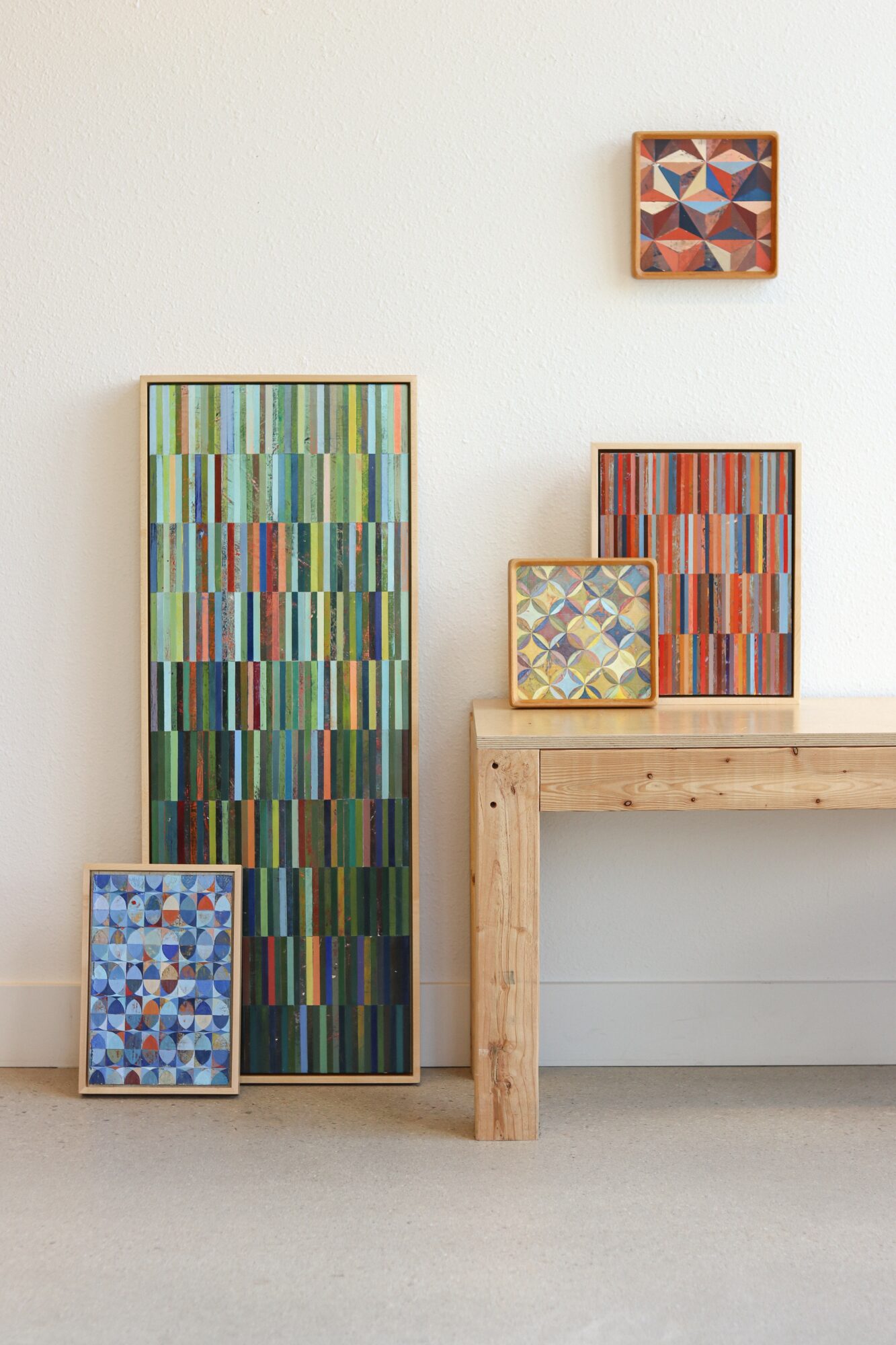 Display of five colorful abstract paintings on a white wall and wooden table, with a light-colored floor.