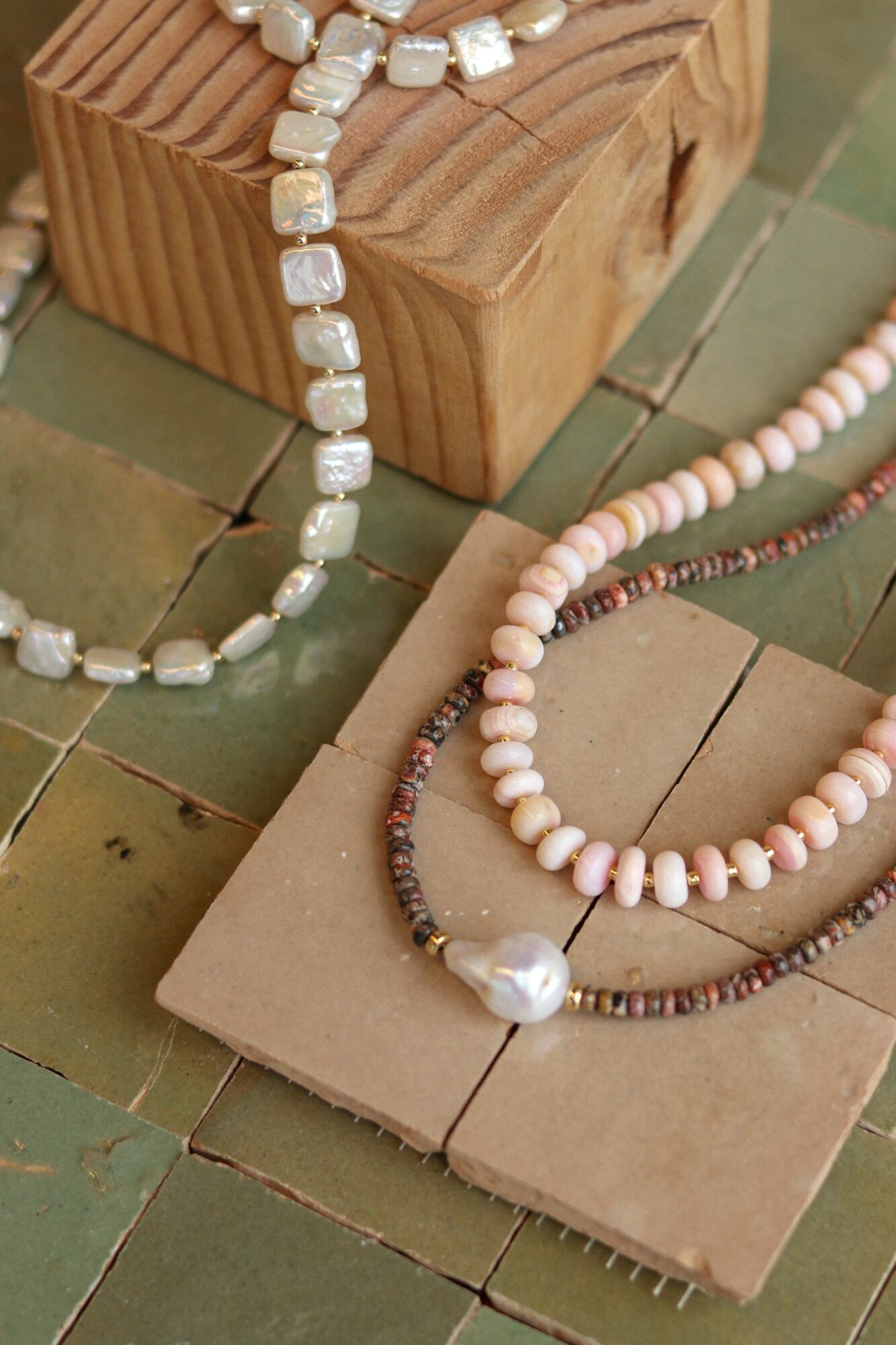 Three necklaces with beads and a large pearl, displayed on a tiled surface and wooden block.