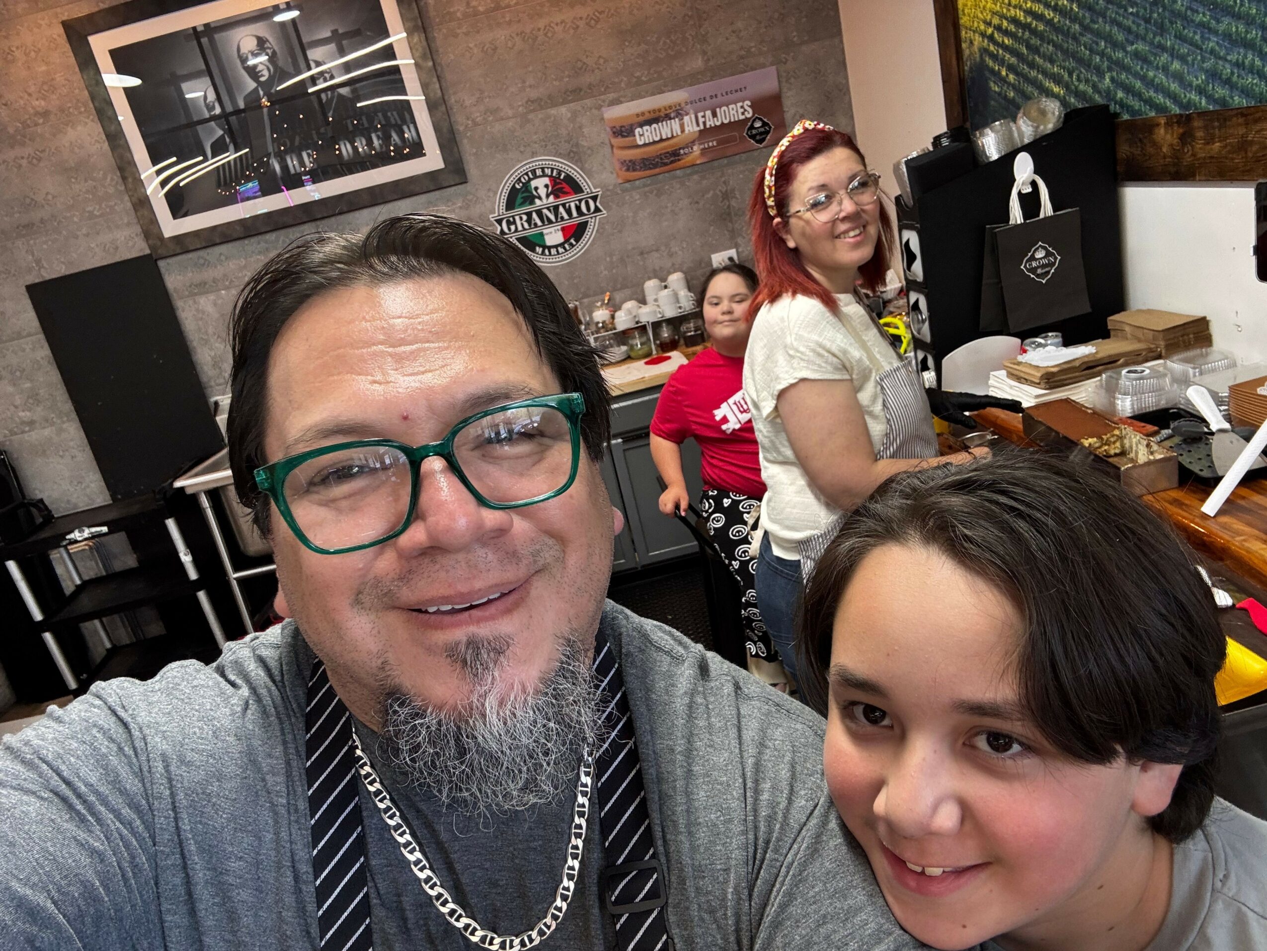Two people taking a selfie indoors, with a woman in the background and a man in the foreground wearing glasses.