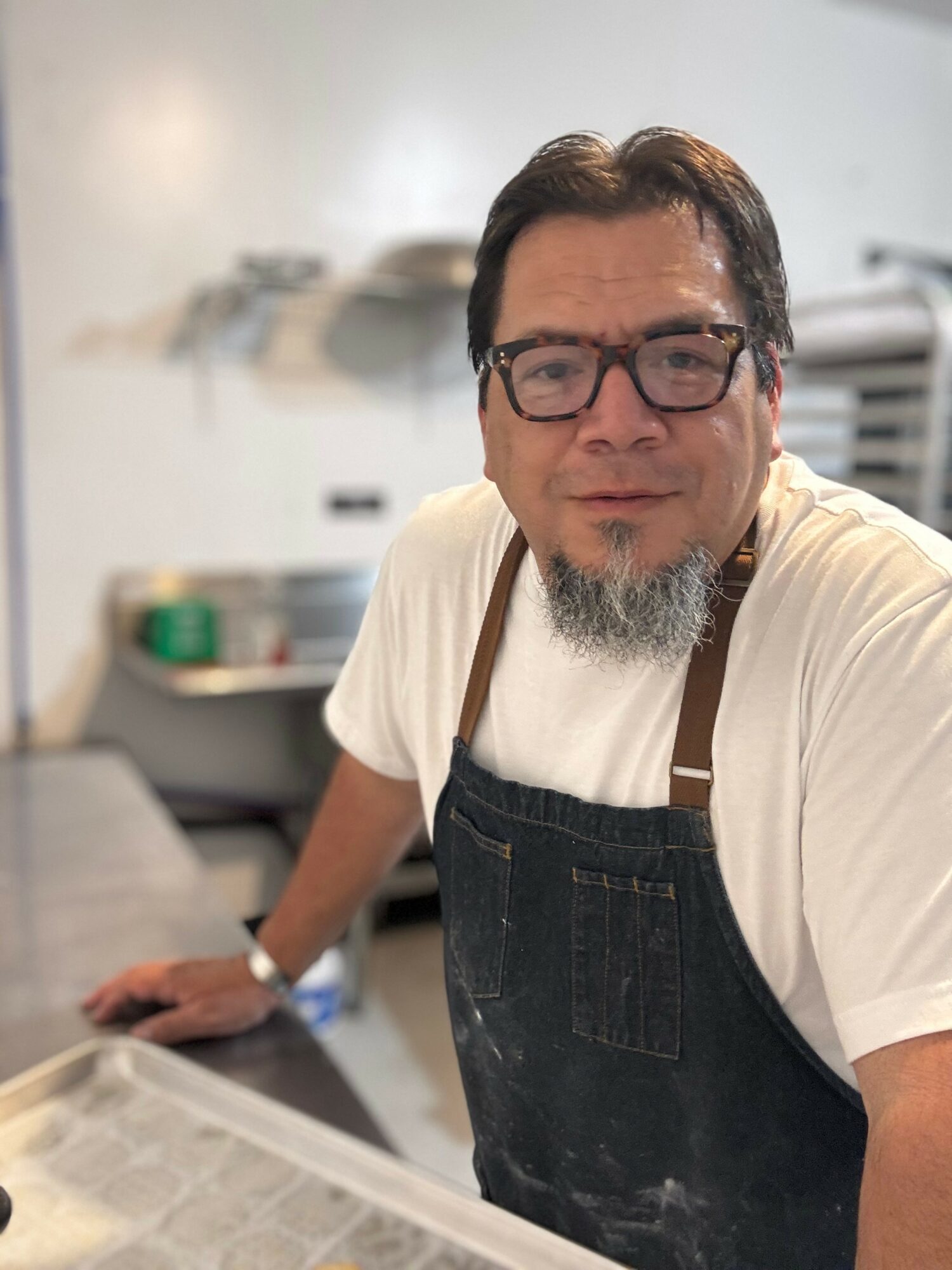 Man with glasses and a beard wearing a white shirt and apron in a kitchen, smiling at the camera.
