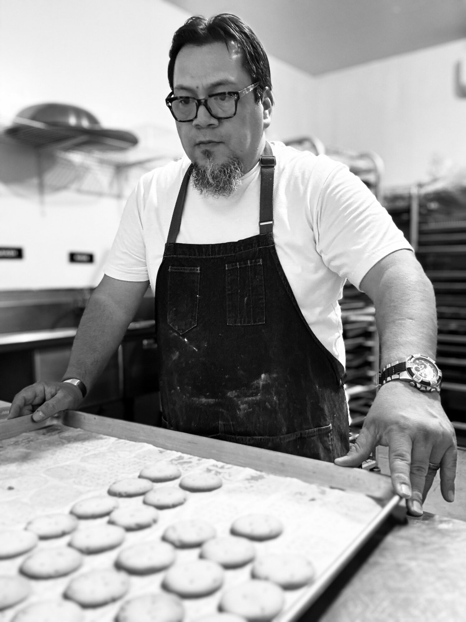 Man wearing glasses and apron, preparing cookies on a baking tray in a kitchen.