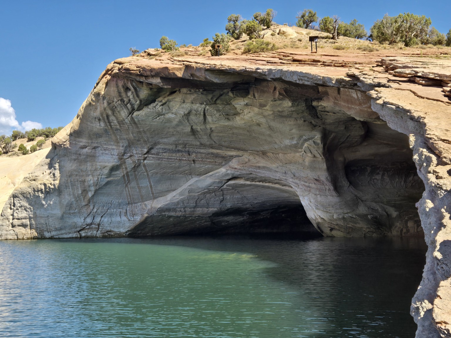 Cliff with a large cave opening over water, blue sky, and some vegetation on top.