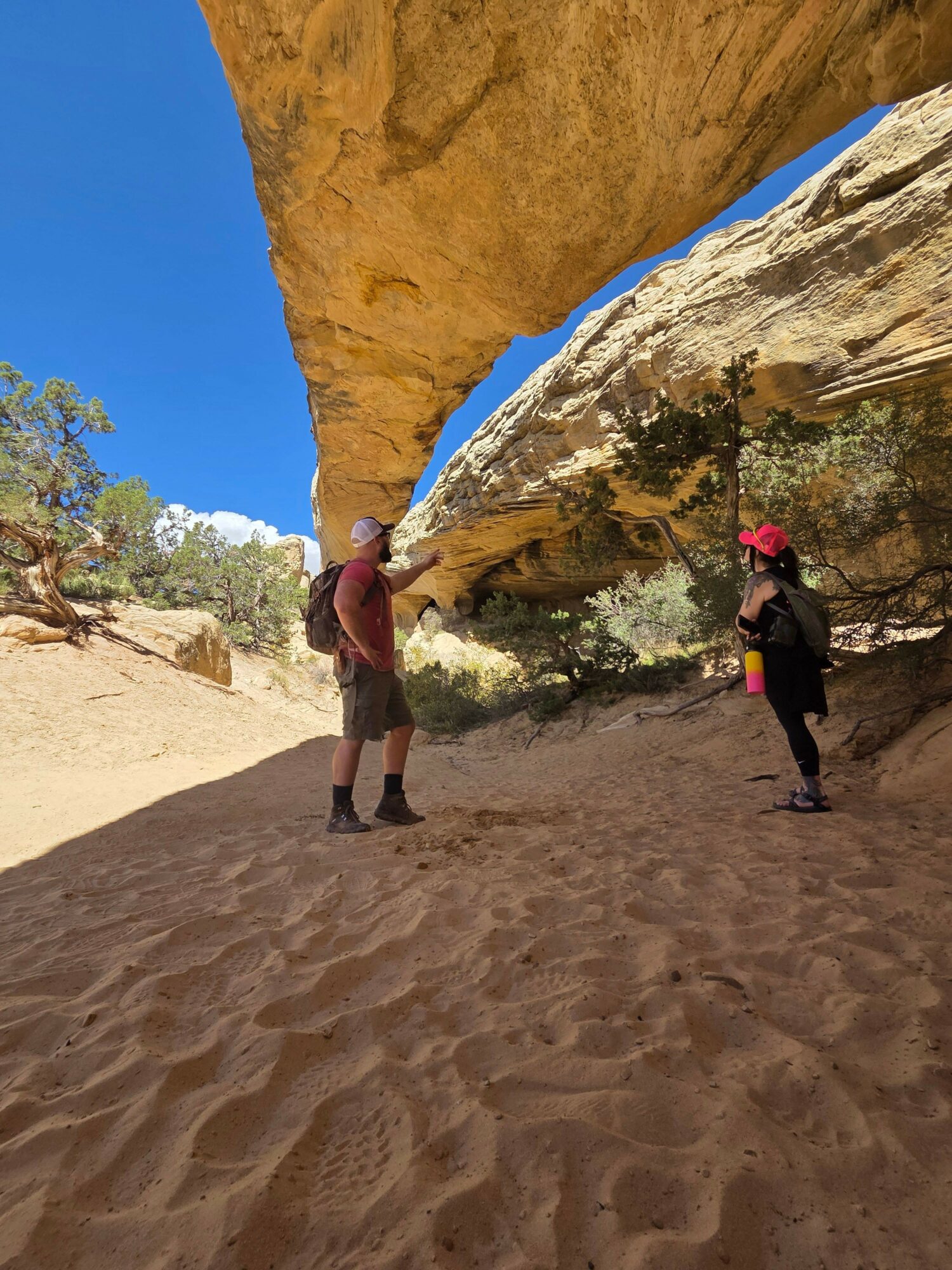Two people walking on sandy terrain near tall rock formations with a clear blue sky.