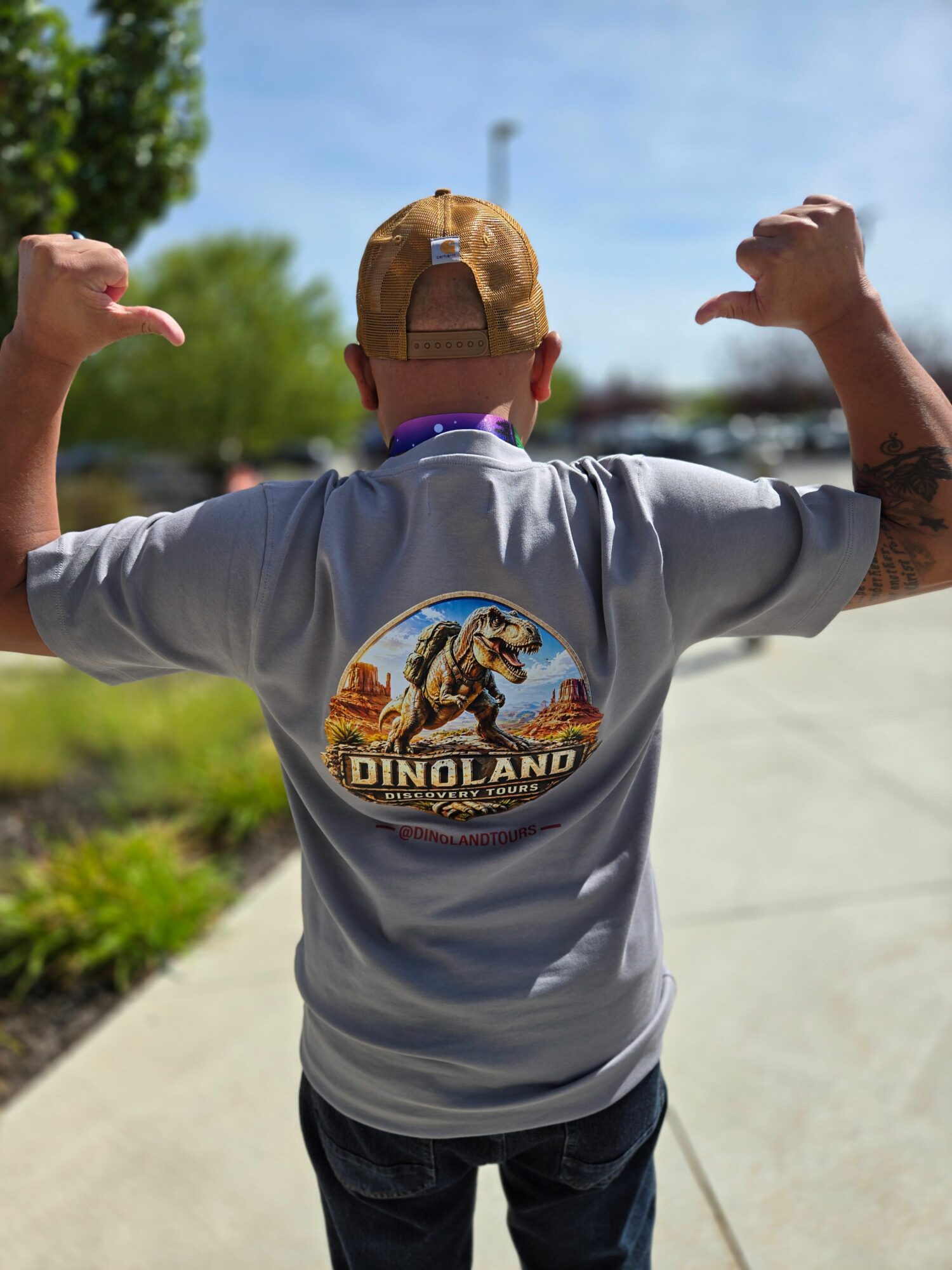 Child wearing a gray T-shirt with a dinosaur graphic, standing outdoors with trees and sky in background.