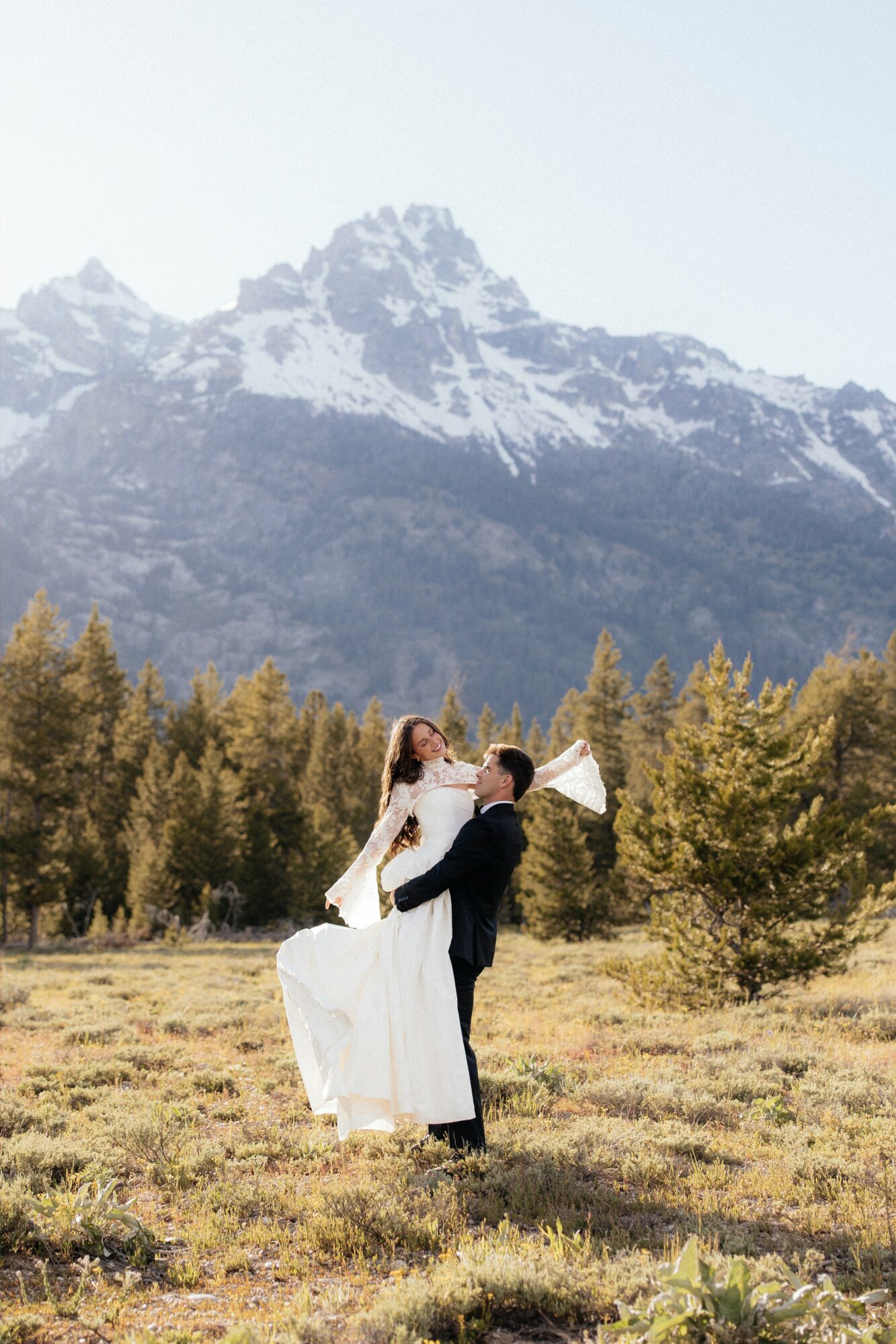 A couple in wedding attire standing in a field with mountains and trees in the background.